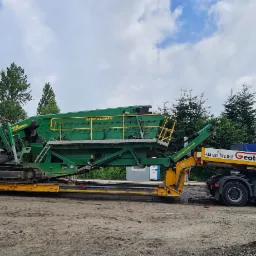 Duża, zielona przesiewaczka mobilna 5x20 SCREEN na żółtej lawecie, transportowana przez białą ciężarówkę GEOTAR, obok biały samochód dostawczy z otwartym bagażnikiem, na tle drzew i pochmurnego nieba.