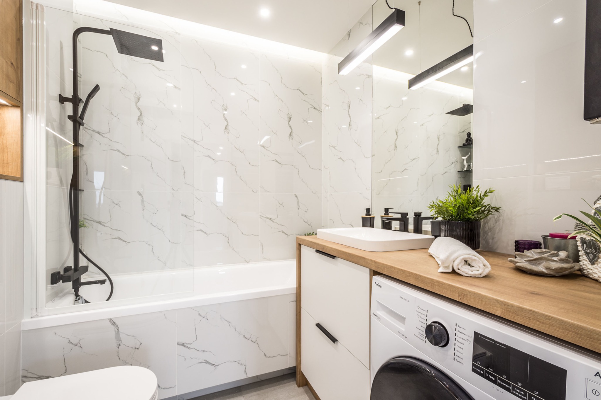 Nowoczesna łazienka z wanną, kabiną prysznicową typu walk-in, umywalką na drewnianym blacie, pralką i marmurowymi płytkami na ścianach, oświetlona podłużną lampą sufitową.