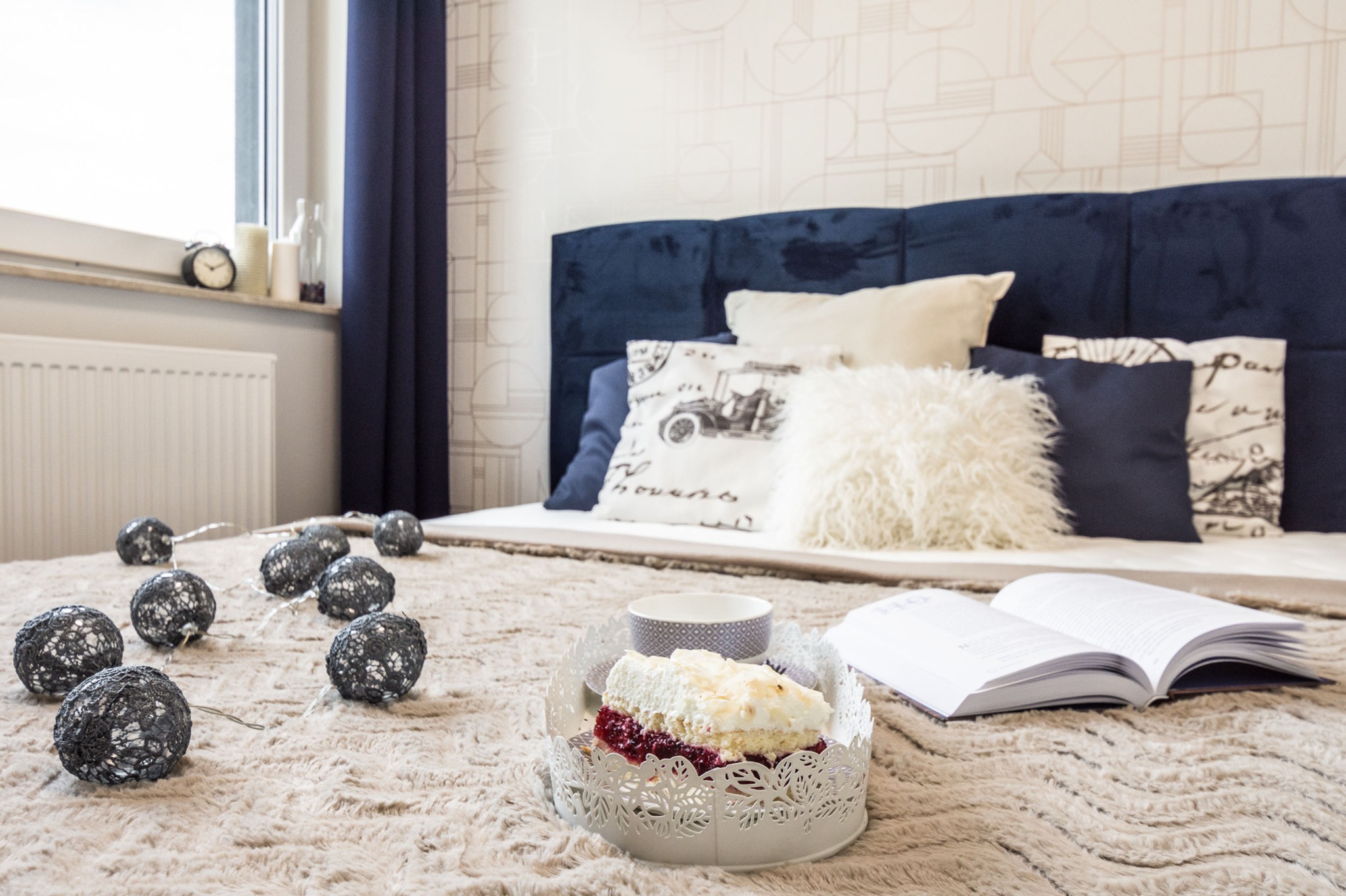 Przytulne wnętrze sypialni z niebieskim zagłówkiem, dekoracyjnymi poduszkami, lampkami w kształcie kul, książką i ciastem na łóżku.