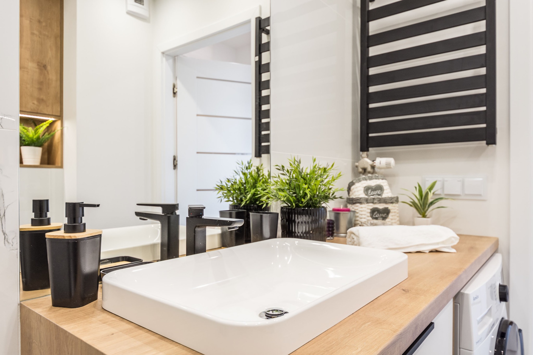 Nowoczesna łazienka z białą umywalką nablatową, czarną armaturą i drewnianym blatem, uzupełniona o dekoracyjne rośliny doniczkowe i grzejnik drabinkowy.