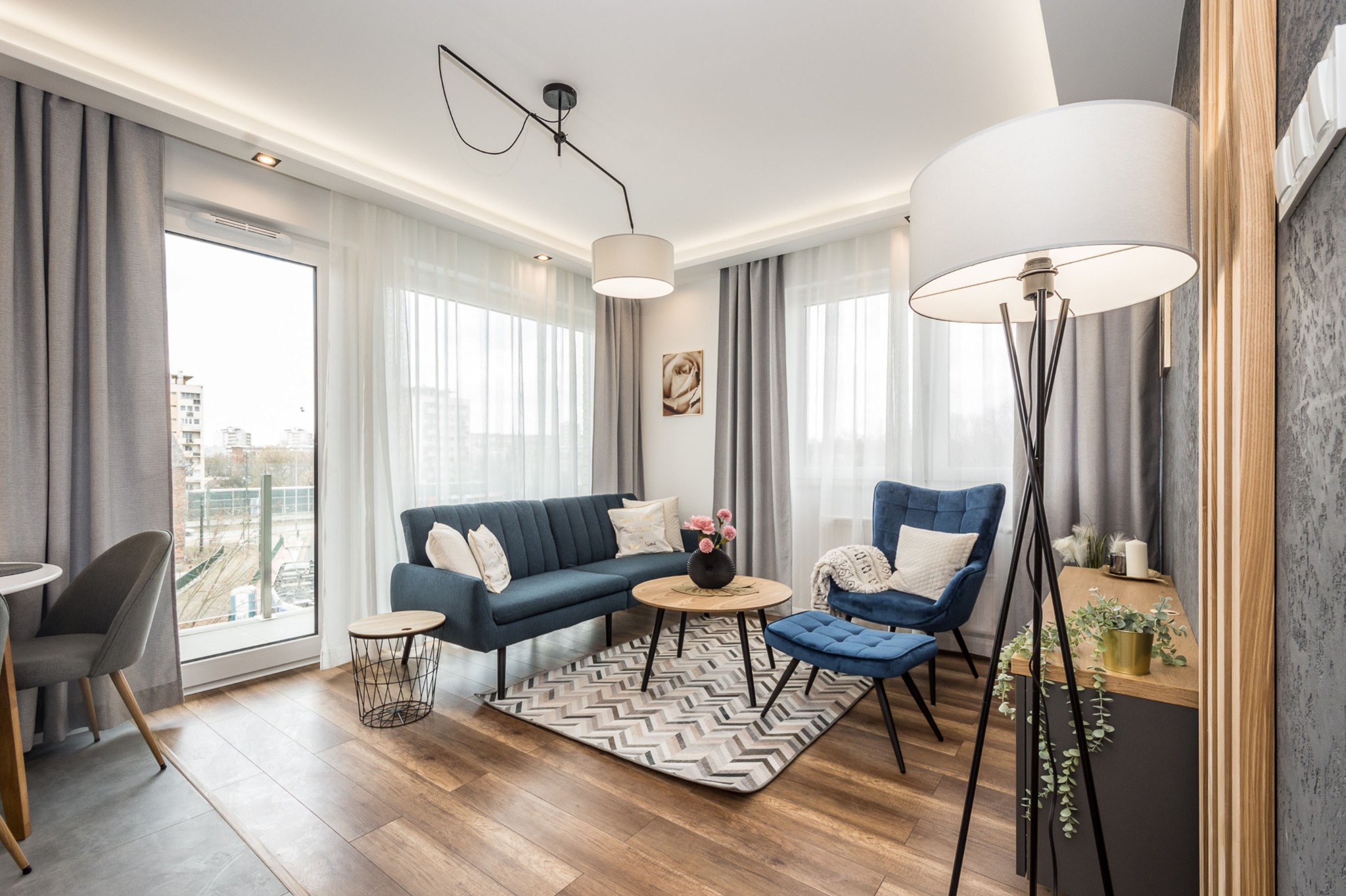 Nowoczesny salon z niebieską sofą, fotelem i pufą na geometrycznym dywanie, oświetlony lampą podłogową i wiszącą, z widokiem na miasto przez okno.