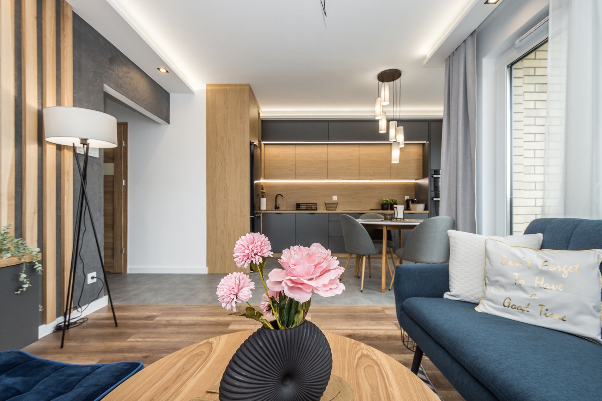 Nowoczesne wnętrze mieszkania: salon z kuchnią, drewniana podłoga, niebieska sofa z dekoracyjnymi poduszkami, okrągły stolik kawowy z czarnym wazonem i różowymi kwiatami, oświetlenie LED...