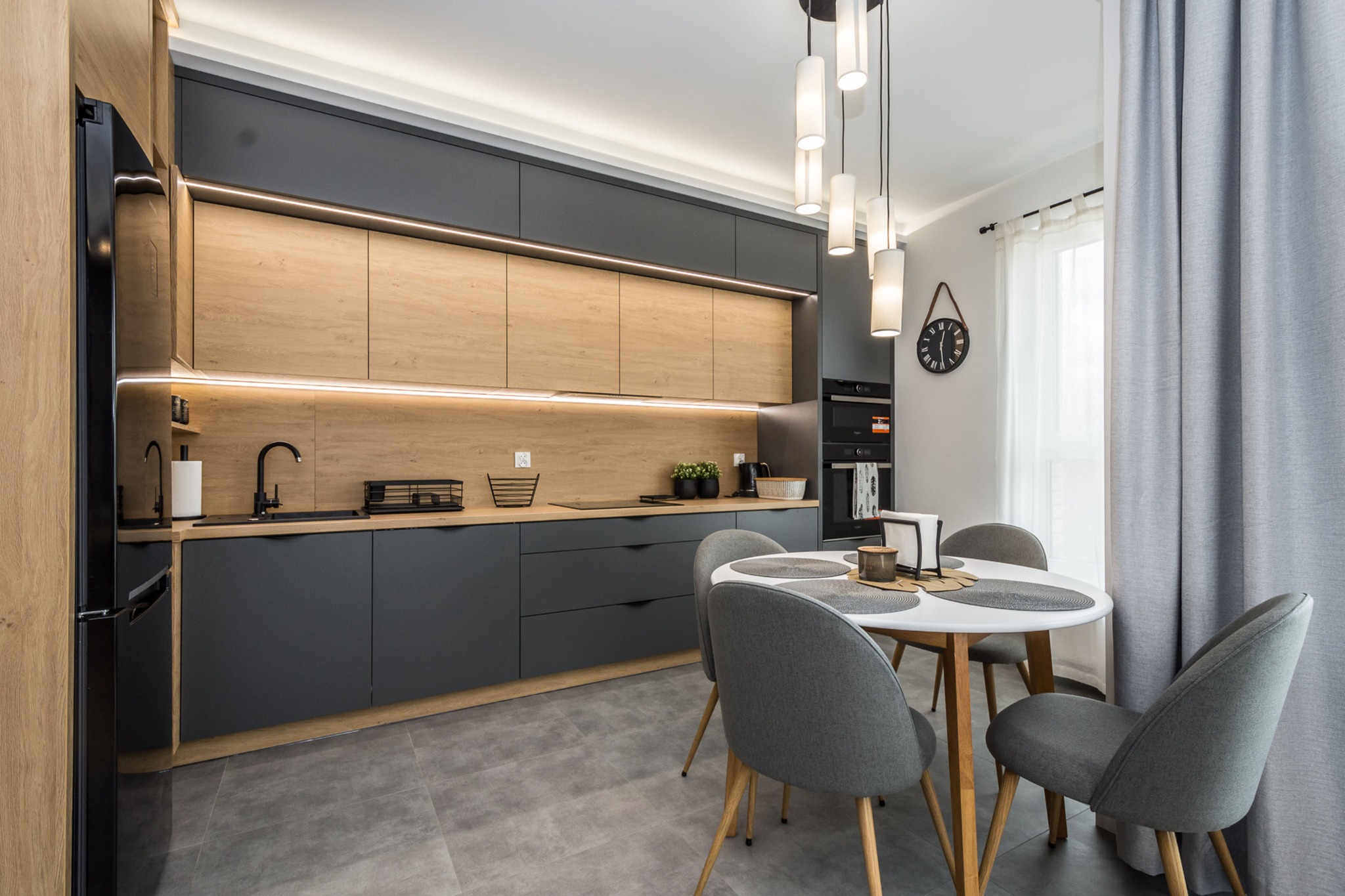 Nowoczesna kuchnia z szarymi szafkami, drewnianymi akcentami i okrągłym stołem z szarymi krzesłami, oświetlona wiszącymi lampami i listwami LED.
