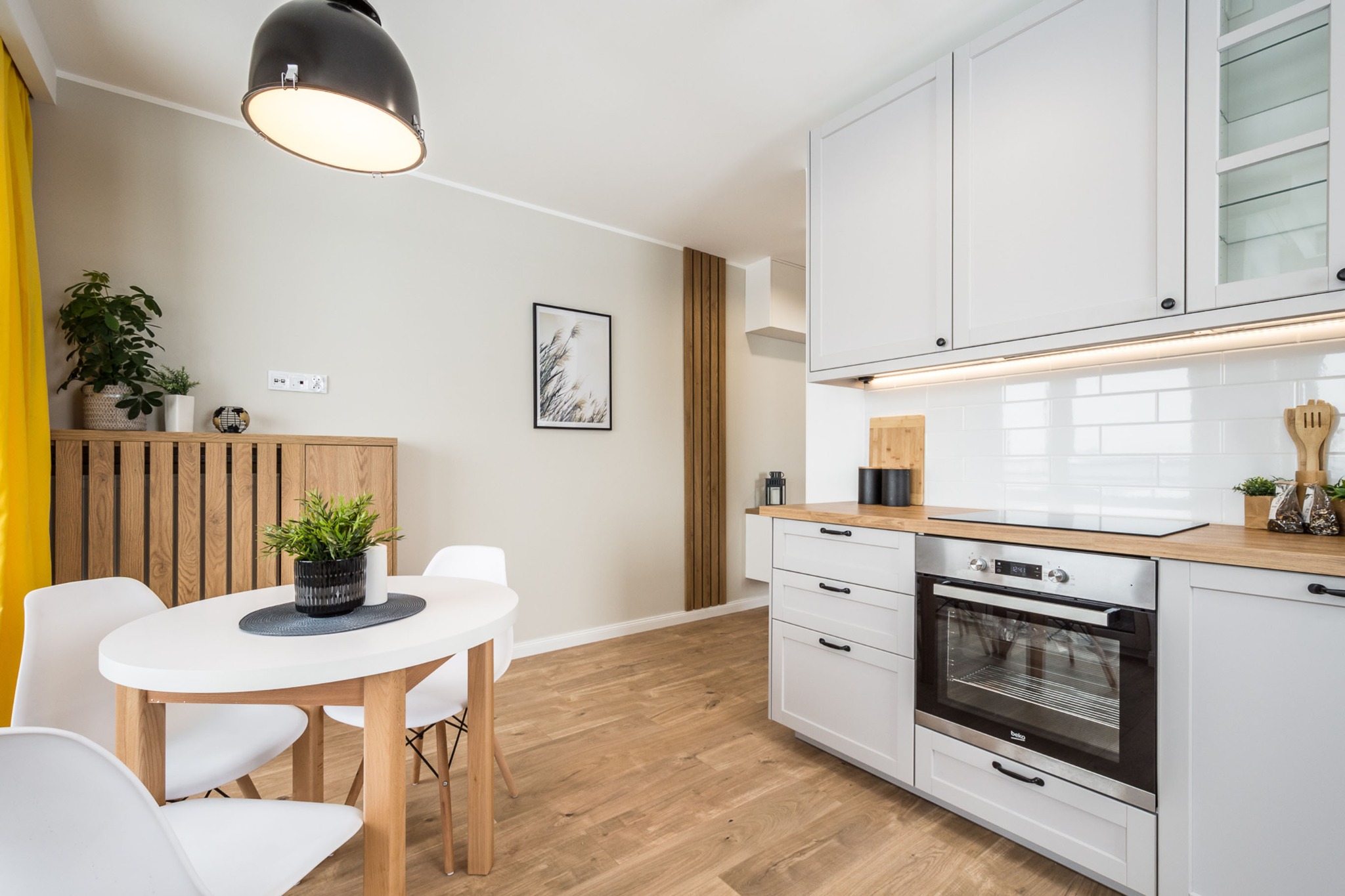 Jasna kuchnia z białymi szafkami, drewnianym blatem i okrągłym stołem z białymi krzesłami, oświetlona lampą w stylu industrialnym.