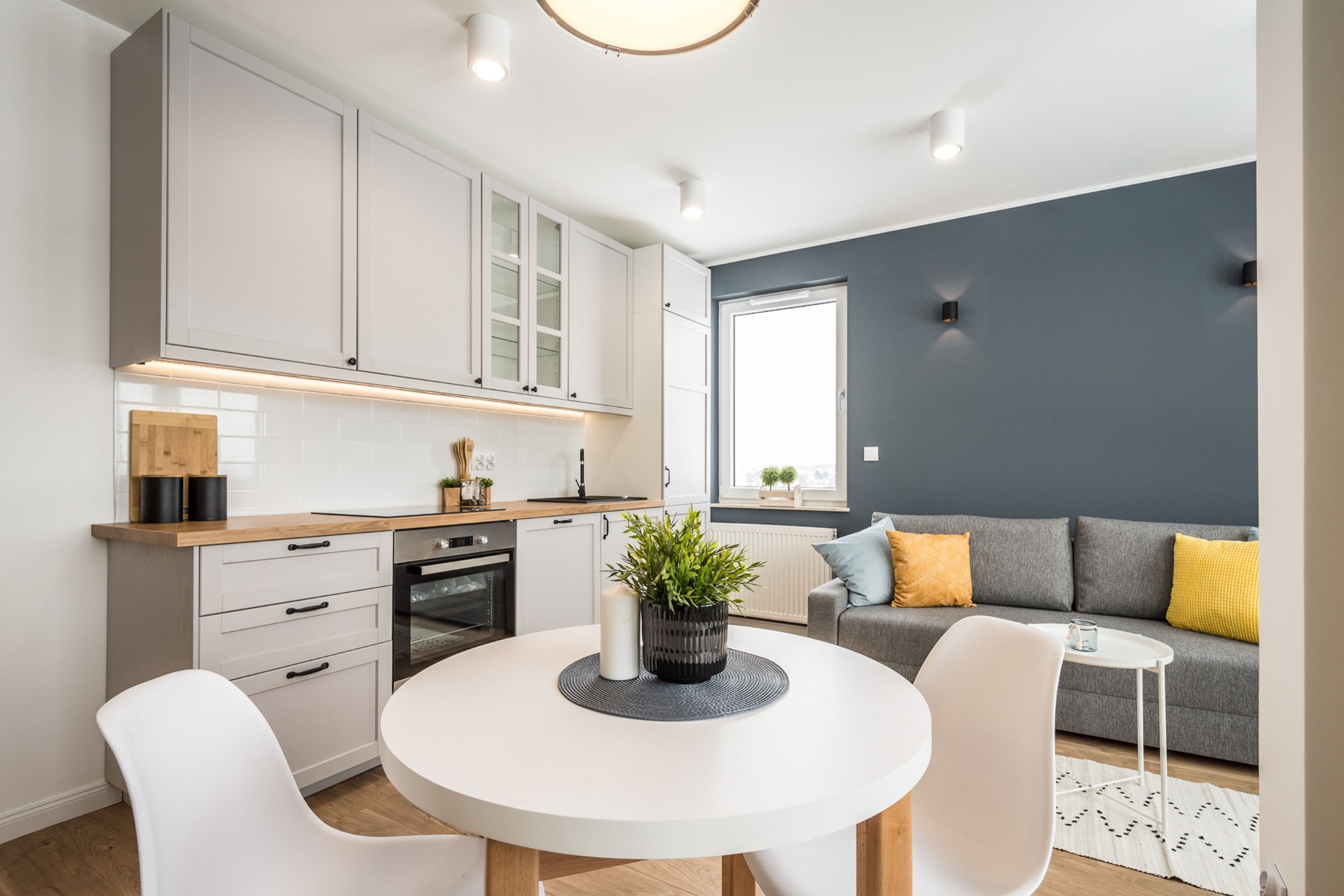 Jasne, nowoczesne wnętrze mieszkania z aneksem kuchennym, okrągłym stołem i szarą sofą z kolorowymi poduszkami.