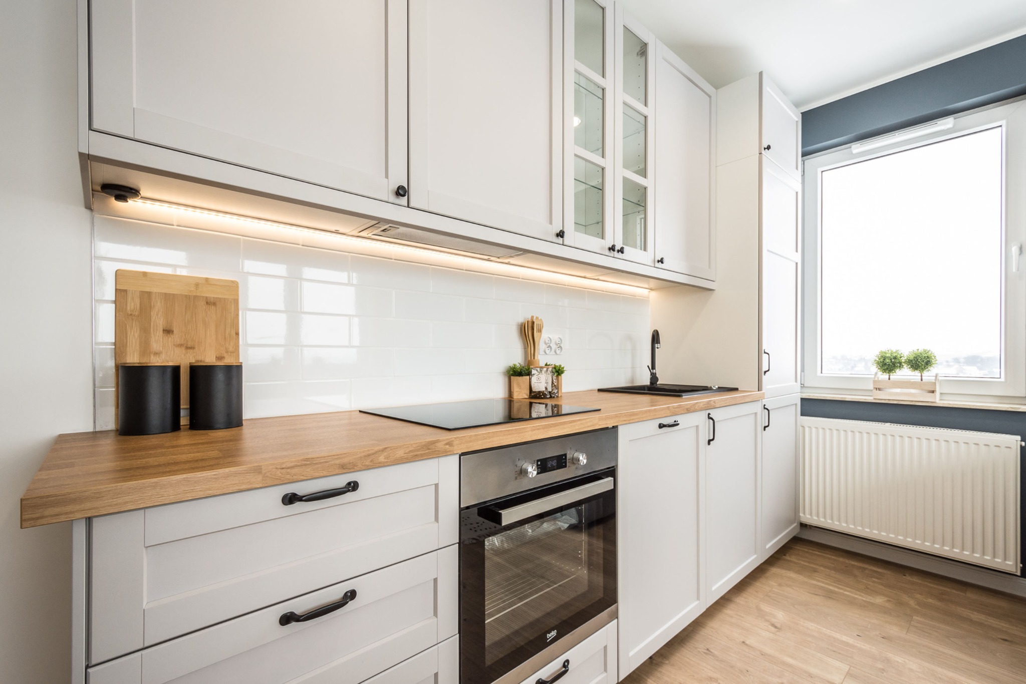 Jasna kuchnia z drewnianym blatem, białymi szafkami z ciemnymi uchwytami, płytą indukcyjną i piekarnikiem, oświetlona taśmą LED pod szafkami wiszącymi.
