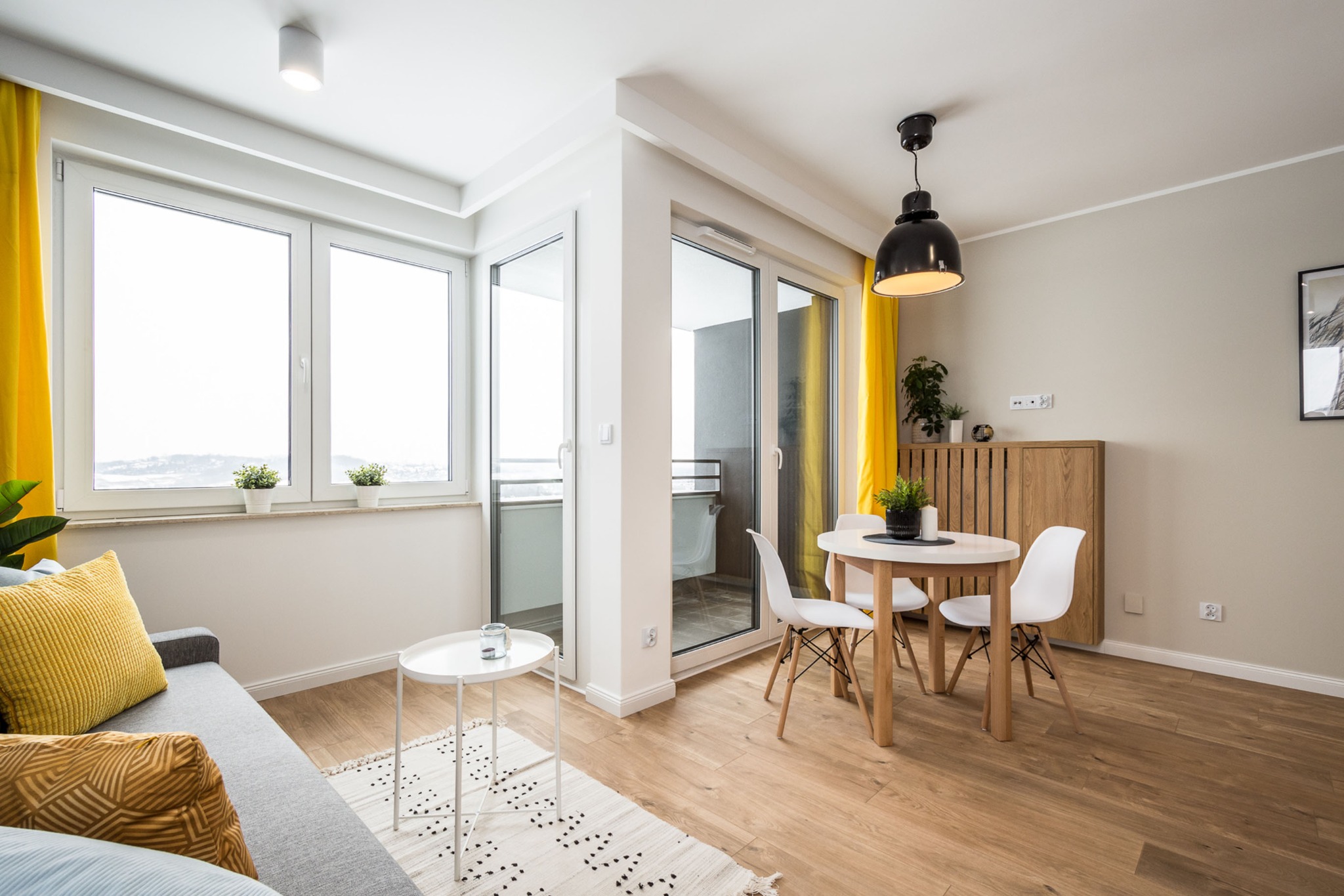 Jasne wnętrze mieszkania z drewnianą podłogą, okrągłym stołem z białymi krzesłami, szarą sofą z żółtymi poduszkami i żółtymi zasłonami w oknach.