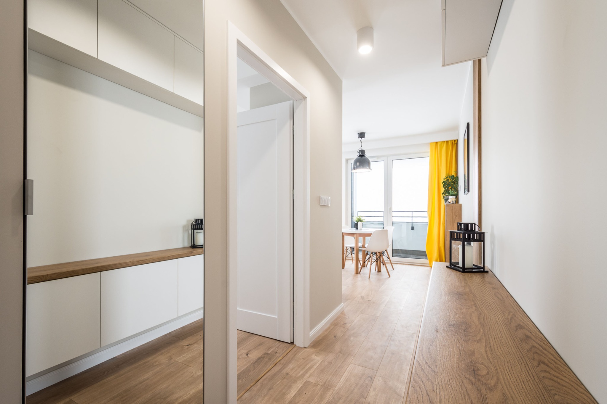 Nowoczesny przedpokój z drewnianą podłogą, białymi ścianami i szafkami, lustrem oraz widokiem na salon z żółtymi zasłonami i stołem.