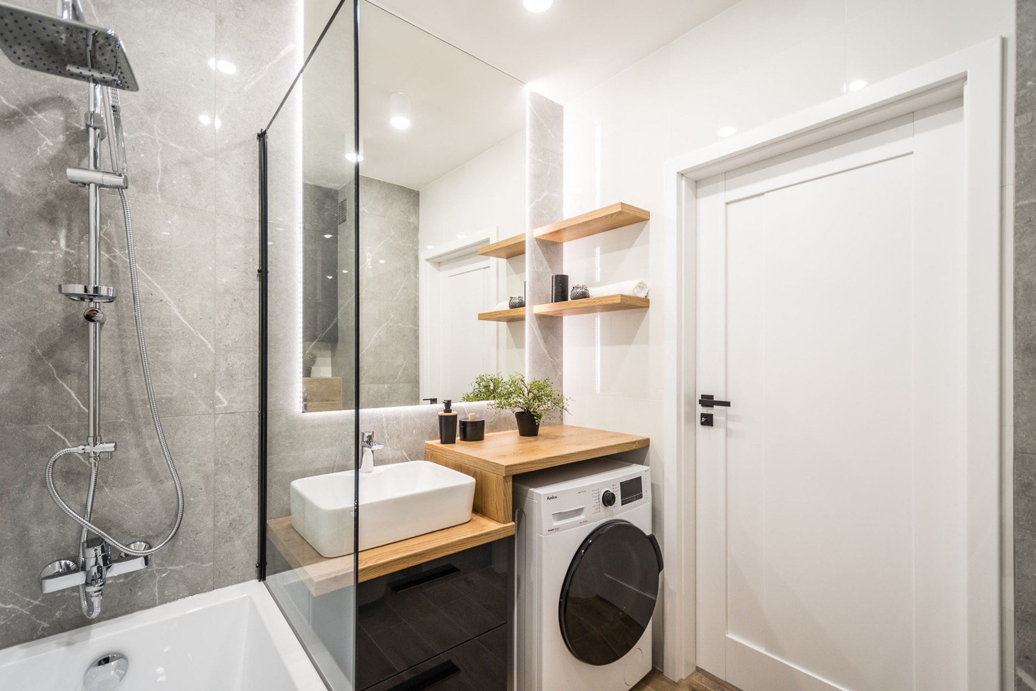 Nowoczesna łazienka z marmurowymi płytkami, kabiną prysznicową ze szklaną ścianką, białą umywalką nablatową, pralką pod drewnianym blatem oraz półkami z dekoracjami.