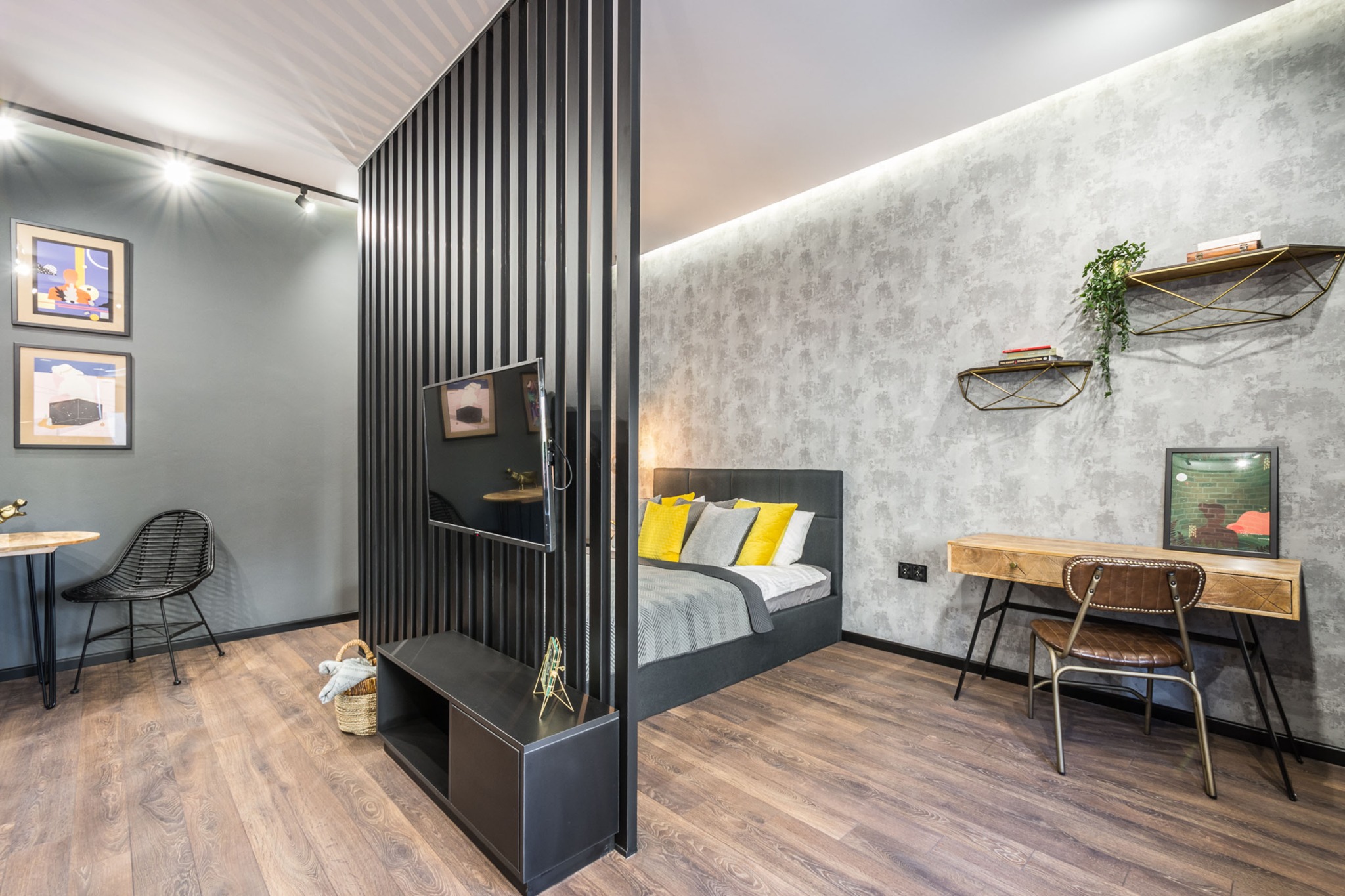 Nowoczesne wnętrze sypialni z drewnianą podłogą, szarą ścianą z półkami i rośliną, oraz łóżkiem z żółtymi poduszkami, oddzielone czarną ażurową ścianką od części z biurkiem i krzesłem.