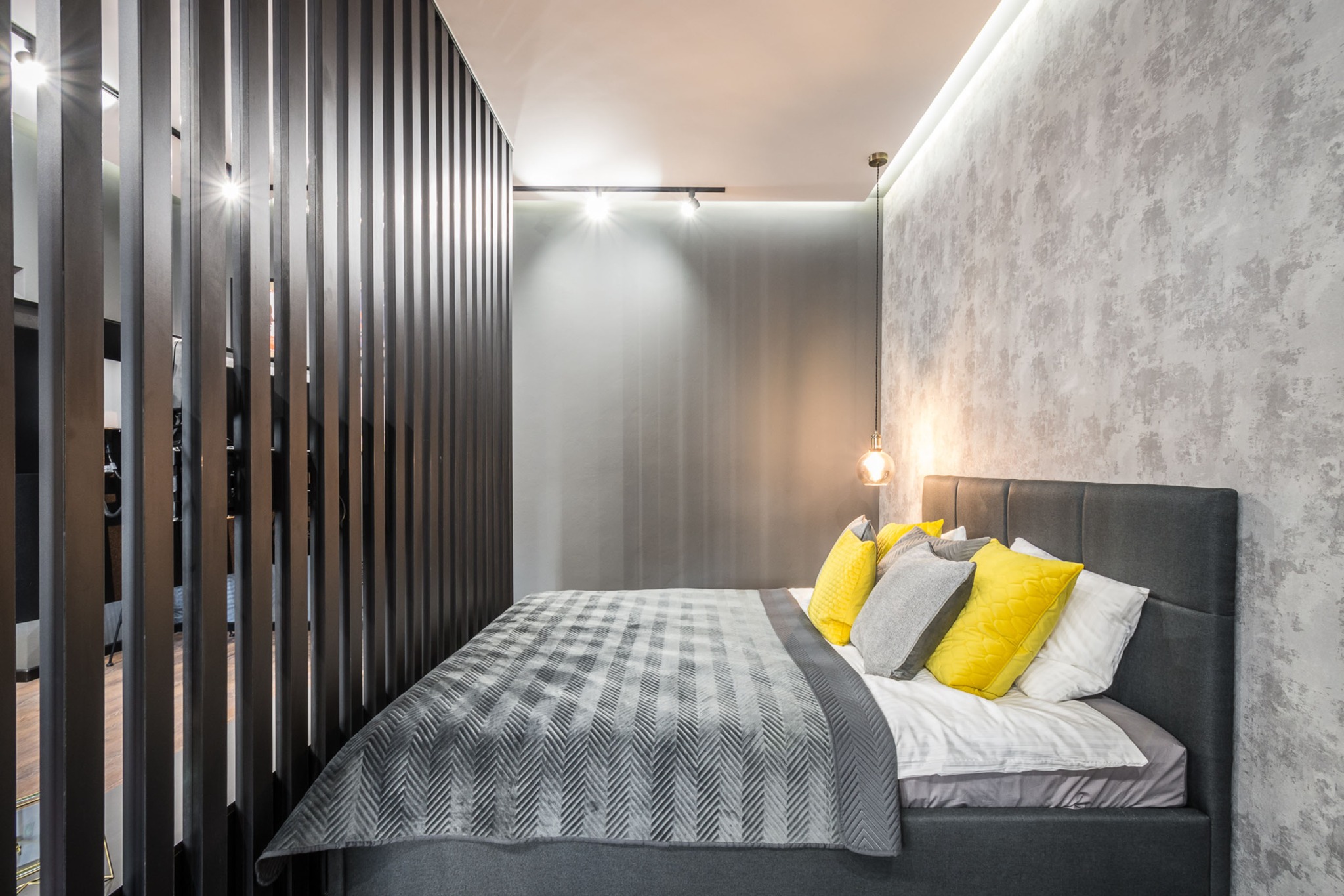 Nowoczesna sypialnia z szarą tapetą imitującą beton, łóżkiem z tapicerowanym zagłówkiem w kolorze grafitu i dekoracyjną ścianką ażurową z pionowych listew.