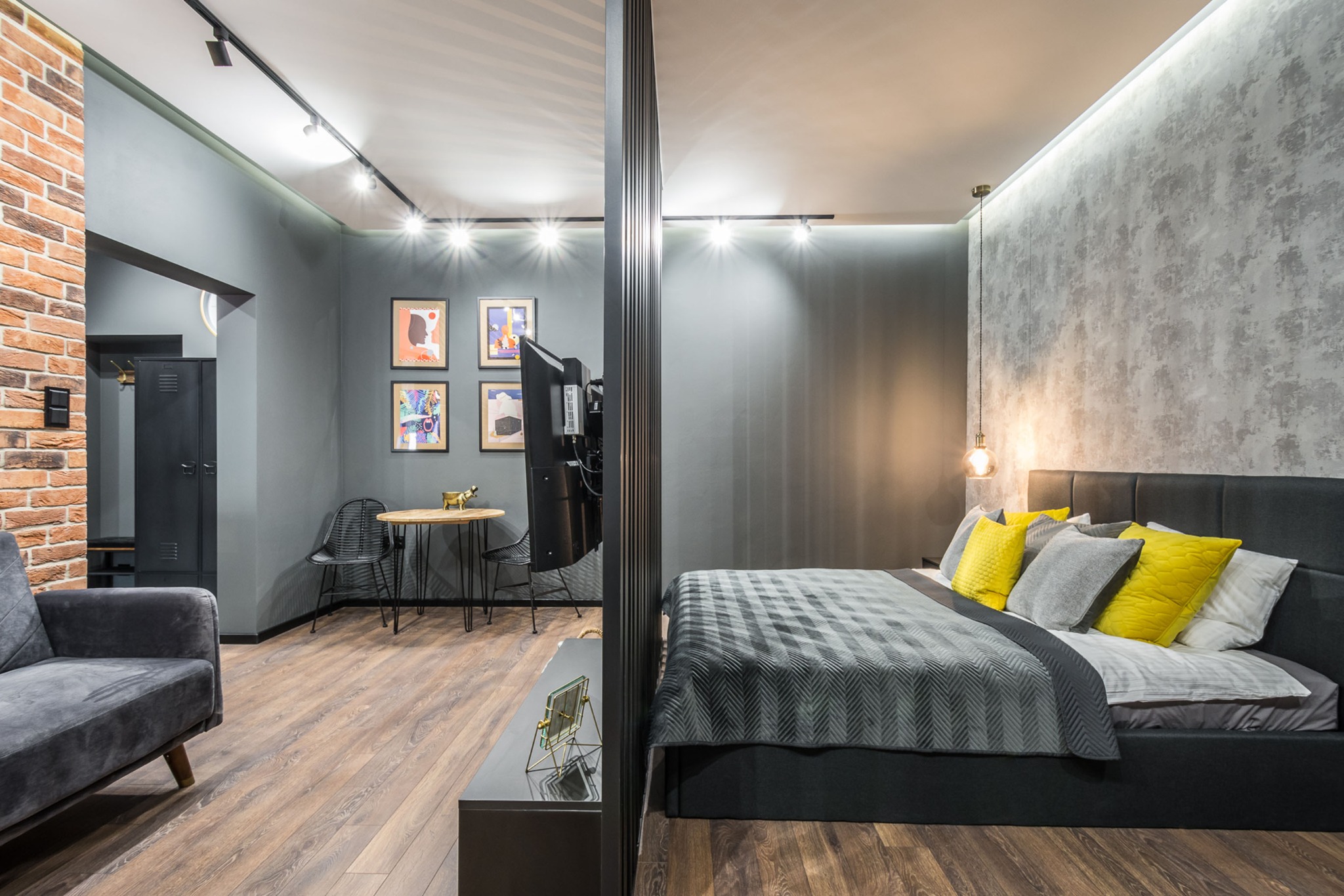 Nowoczesna sypialnia z szarą ścianą akcentową, łóżkiem z szarą tapicerką i żółtymi poduszkami, oddzielona ażurową ścianką od części dziennej z ceglaną ścianą i metalową szafką.