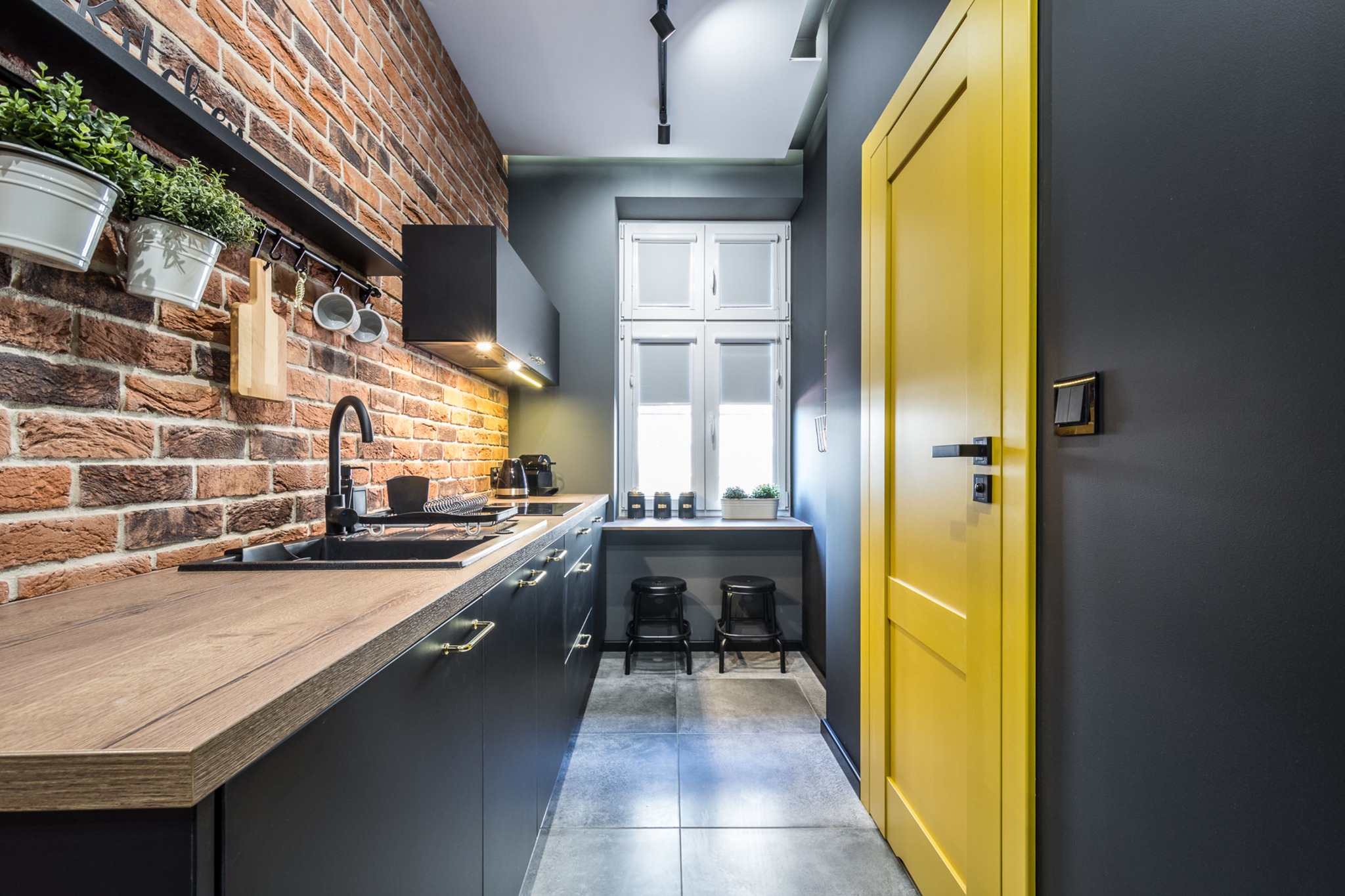Wnętrze kuchni w stylu industrialnym: ceglana ściana, czarne szafki z drewnianym blatem, żółte drzwi i szare ściany. Na ścianie wiszą doniczki z ziołami i akcesoria kuchenne.