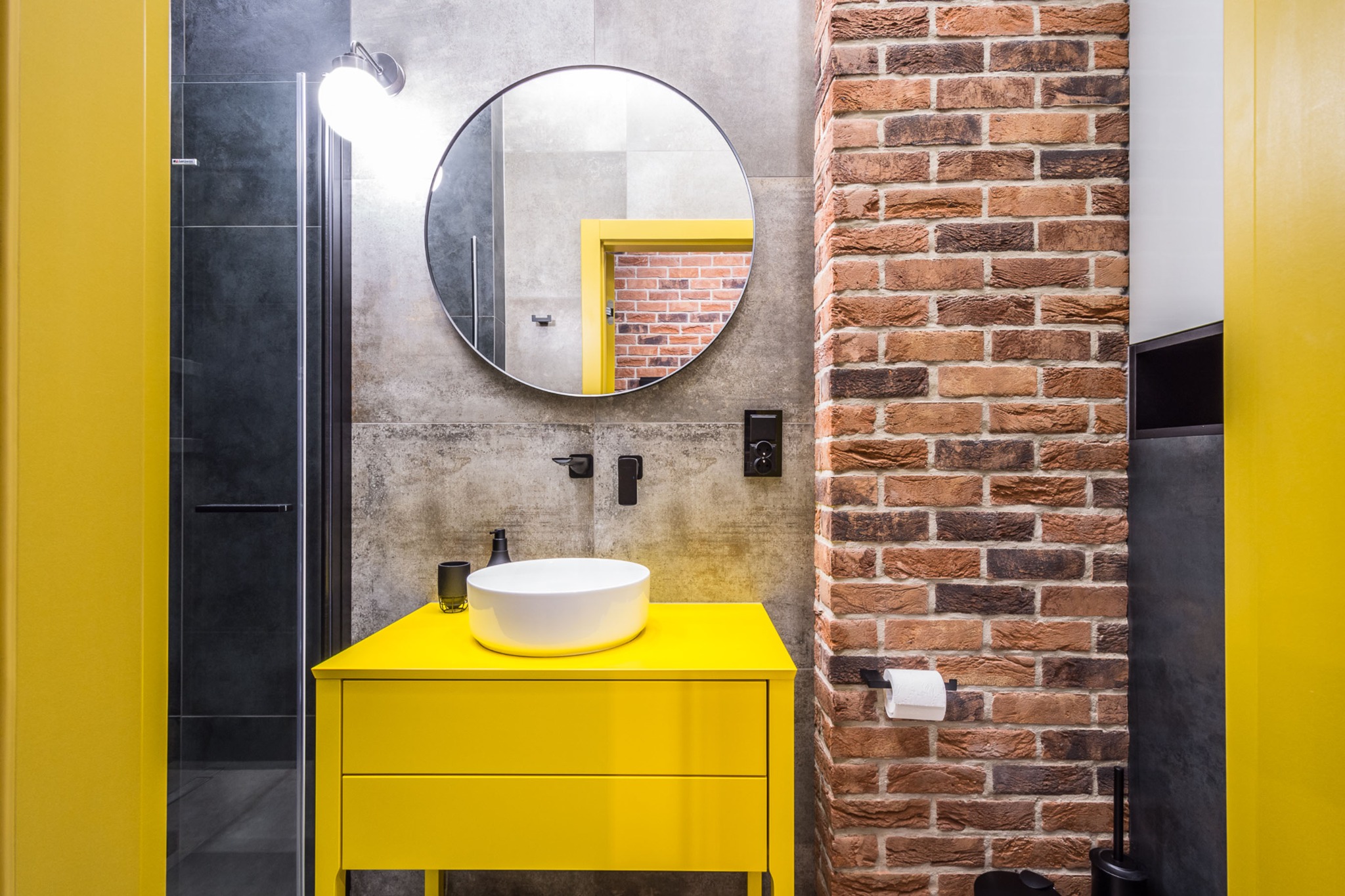 Żółta szafka łazienkowa z białą umywalką na tle ściany z cegły i szarej ściany z okrągłym lustrem, widoczna kabina prysznicowa z czarnymi ramami i żółte obramowanie drzwi w odbiciu lustra.