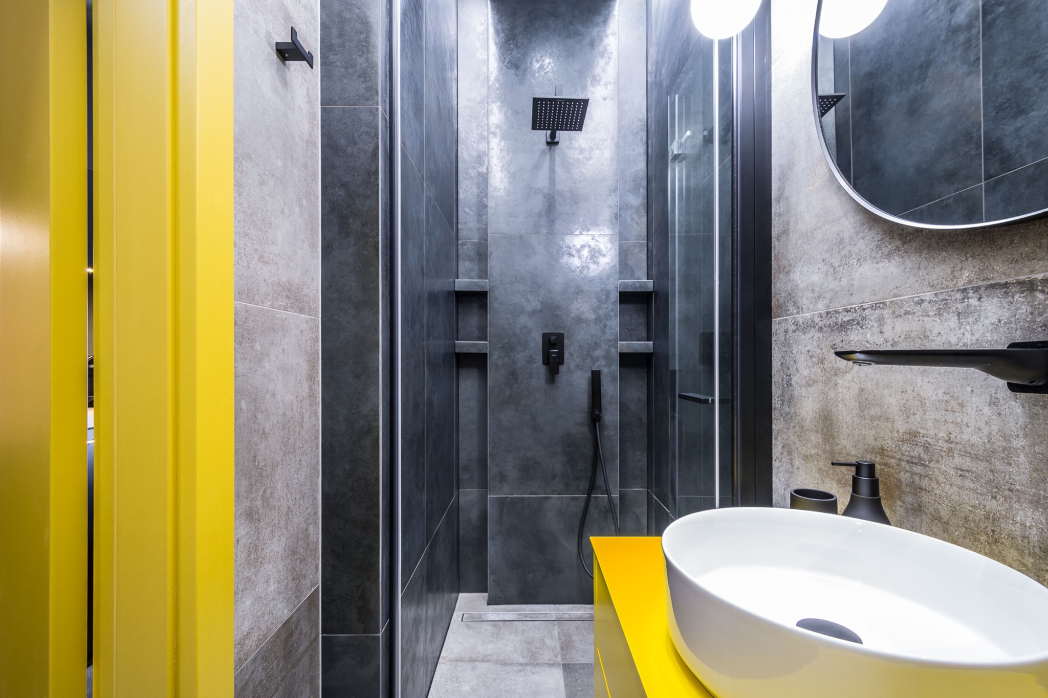 Nowoczesna łazienka z szarą kabiną prysznicową, żółtymi akcentami w postaci drzwi i szafki pod umywalką, okrągłym lustrem i białą umywalką nablatową.