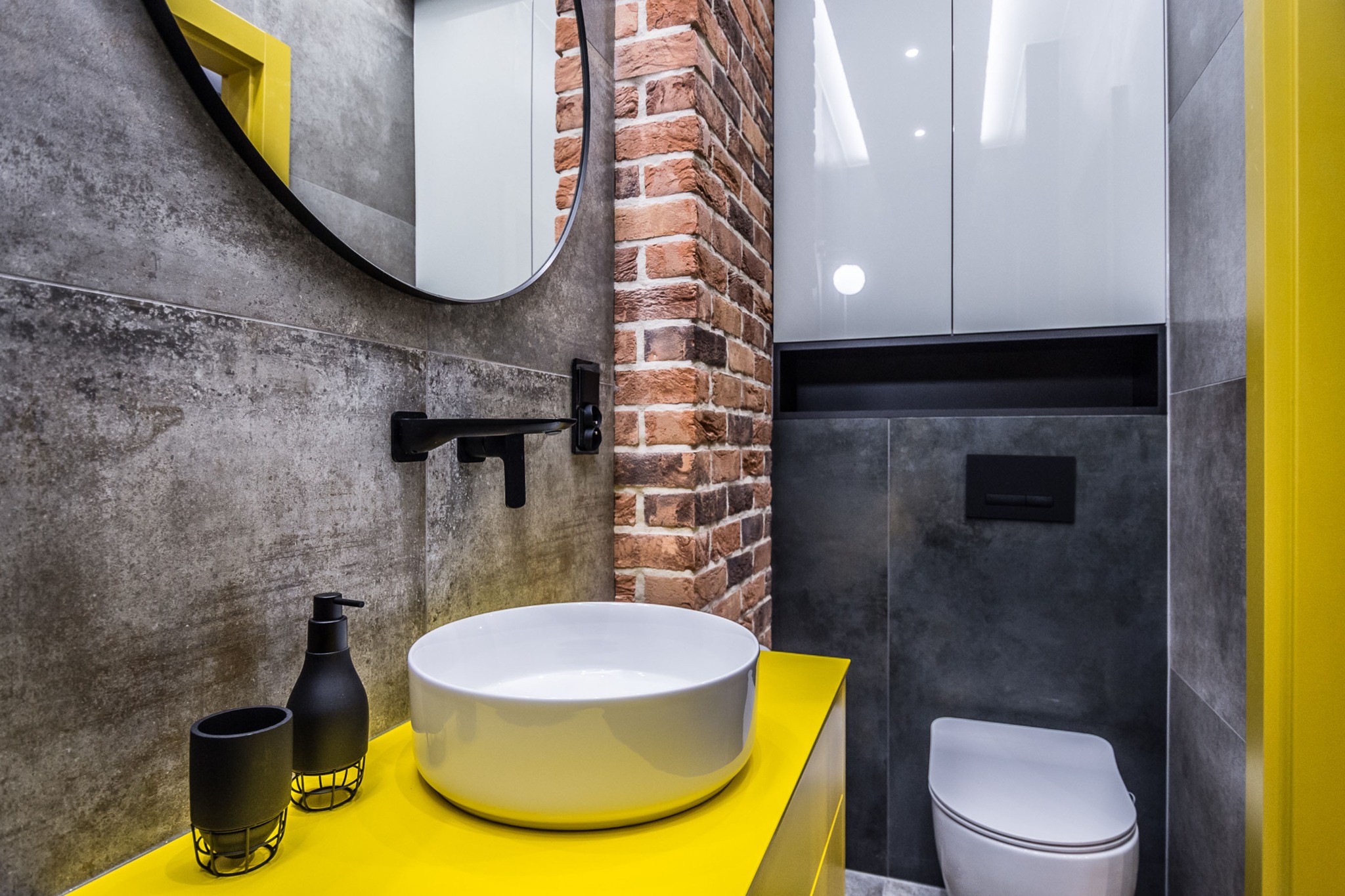 Nowoczesna łazienka w Krakowie z żółtymi akcentami: okrągłe lustro, umywalka nablatowa, ściana z cegły i szara armatura.
