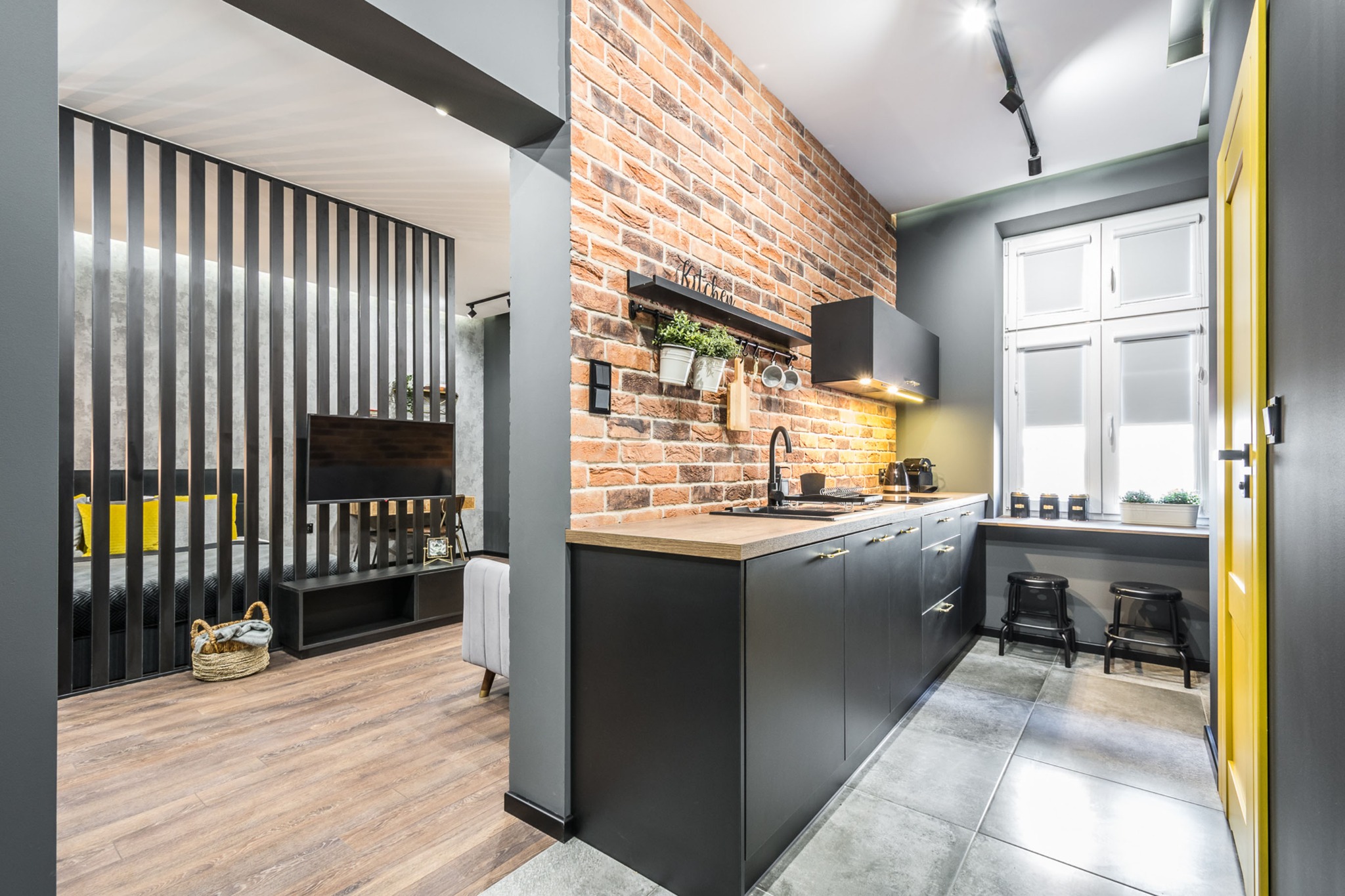 Nowoczesna kuchnia z ceglaną ścianą, czarnymi szafkami i drewnianym blatem, widok na salon oddzielony ażurową ścianką, szare ściany i podłoga, żółte drzwi po prawej.