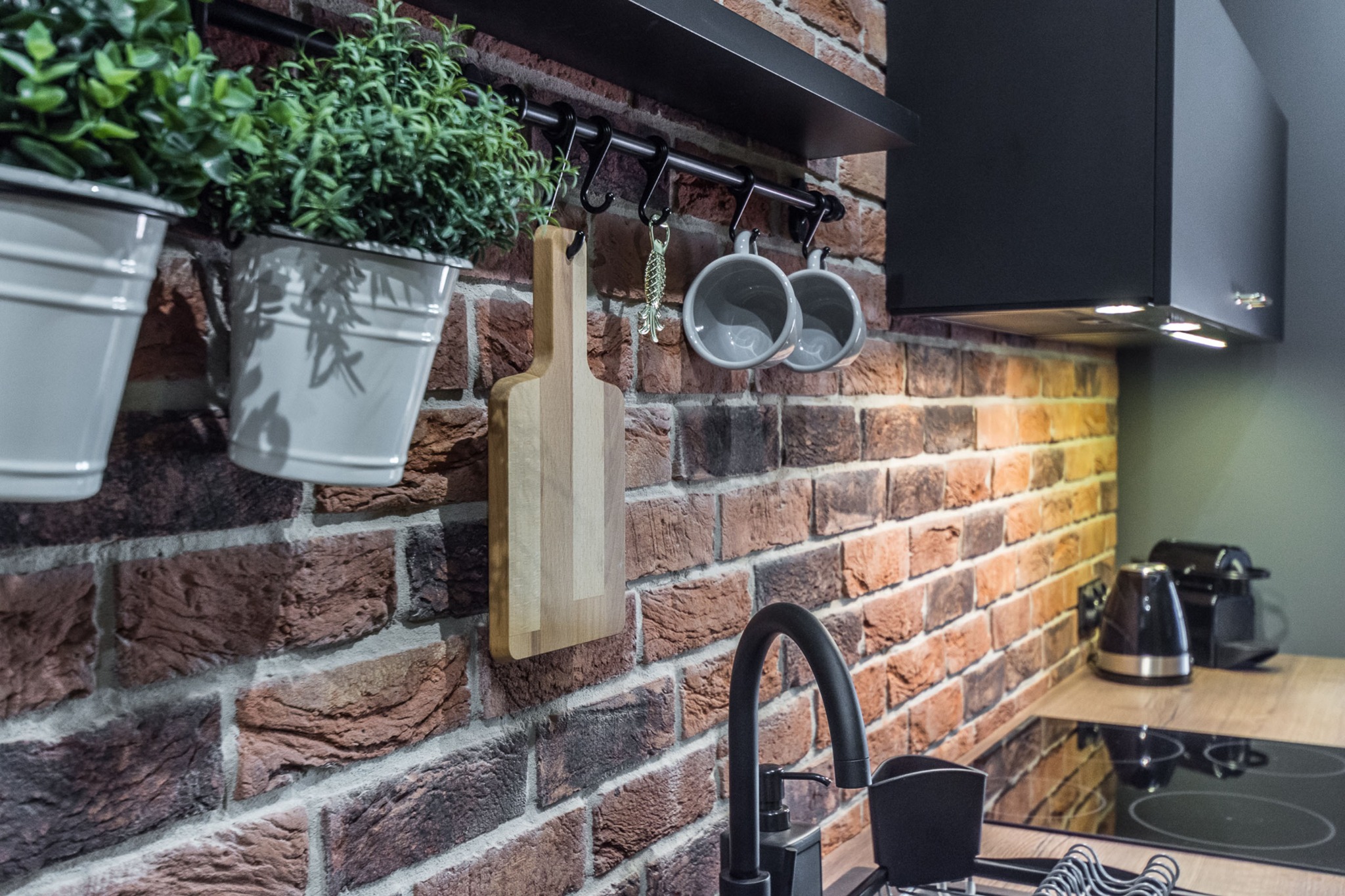 Fragment kuchni z ceglaną ścianą, na której zawieszone są doniczki z ziołami, drewniana deska do krojenia, metalowa zawieszka w kształcie ryby i dwa szare kubki, poniżej bateria kuchenna i widoczny...