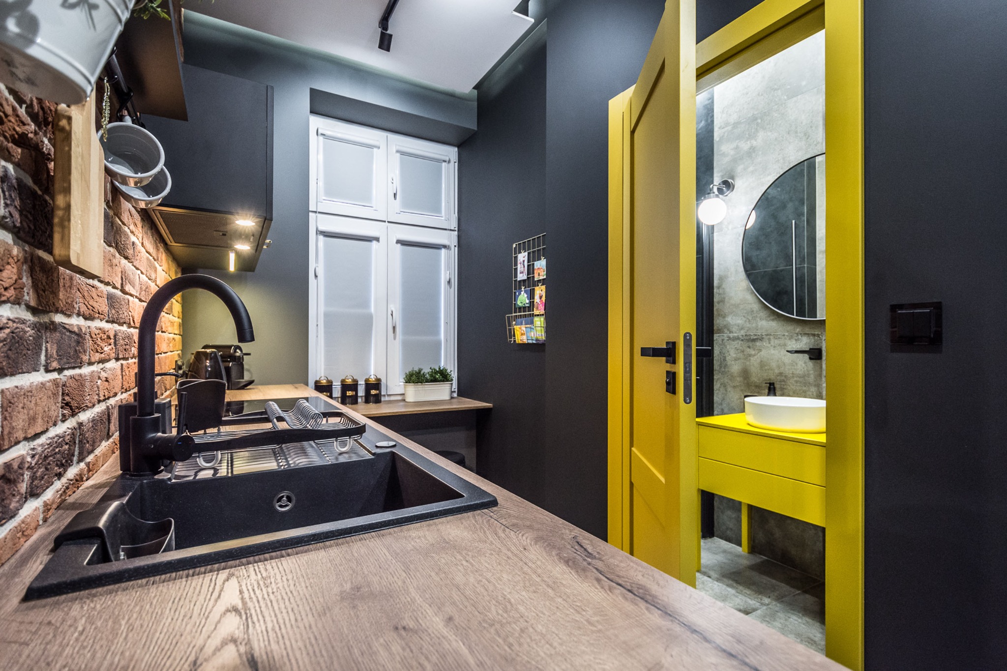 Nowoczesna kuchnia z ceglaną ścianą i czarnym zlewem, widok na łazienkę z żółtymi akcentami przez otwarte drzwi.