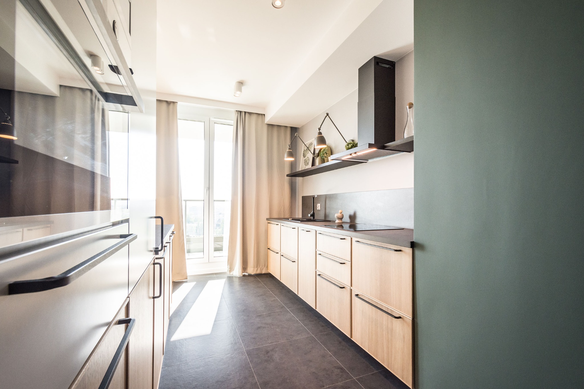 Jasna kuchnia z drewnianymi szafkami, czarnym blatem i podłogą, oświetlona dwoma lampami biurkowymi nad zlewem, z widokiem na balkon przez okno.