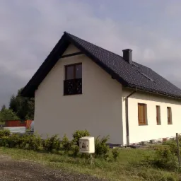 Gotowy dom jednorodzinny z białą elewacją, ciemną dachówką i czarną stolarką okienną, widziany z boku na tle pochmurnego nieba i zieleni.