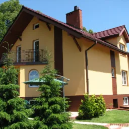 Żółta elewacja domu jednorodzinnego z ciemnobrązowymi akcentami, czerwona dachówka, balkon z ozdobną balustradą, zadbany trawnik z iglakami na pierwszym planie.