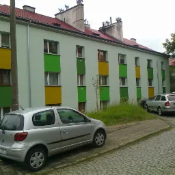 Odnowiony blok mieszkalny z pomalowanymi na zielono i żółto fragmentami elewacji, widok z poziomu ulicy wyłożonej kostką brukową, zaparkowane samochody.