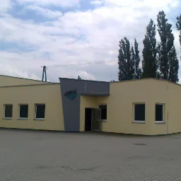 Budynek biurowy z ocieploną elewacją w kolorze kremowym, szarym wejściem i logo firmy, otoczony brukowanym parkingiem i wysokimi drzewami w tle.