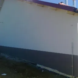 Świeżo otynkowana elewacja budynku z widocznym fragmentem ocieplenia przy gruncie, dwukolorowa z szarą dolną częścią, z brązową rynną i fragmentem dachu w tle.