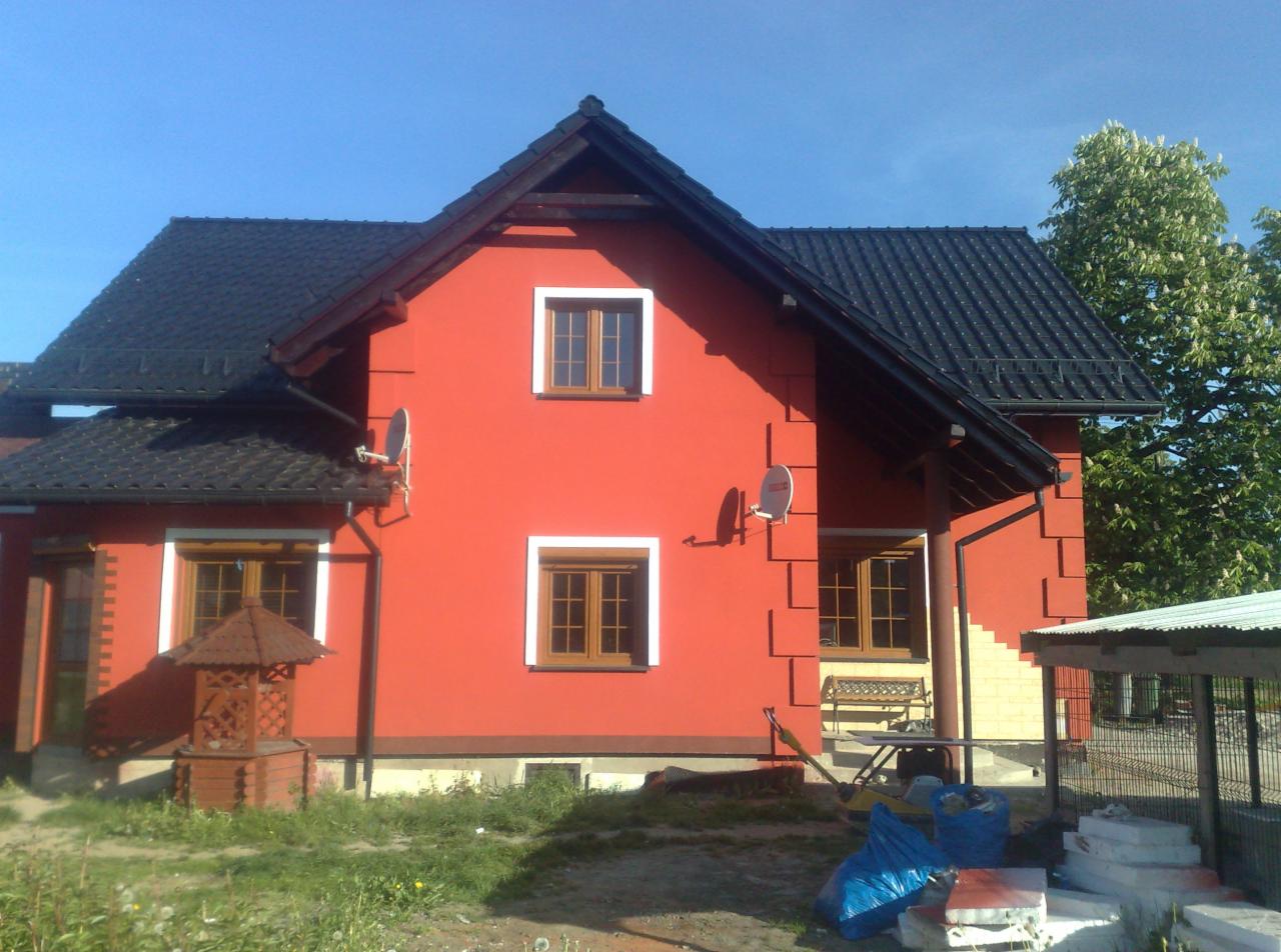 Czerwony dom jednorodzinny z ciemną dachówką, białymi obramowaniami okien i dekoracyjnymi elementami elewacji, widoczne dwie anteny satelitarne, studnia i częściowo odsłonięty fragment ocieplenia...