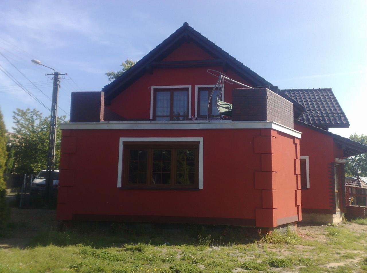 Czerwony dom jednorodzinny z ciemną dachówką, widoczne detale elewacji w postaci wystających bloków oraz huśtawka na balkonie, sfotografowany w słoneczny dzień.