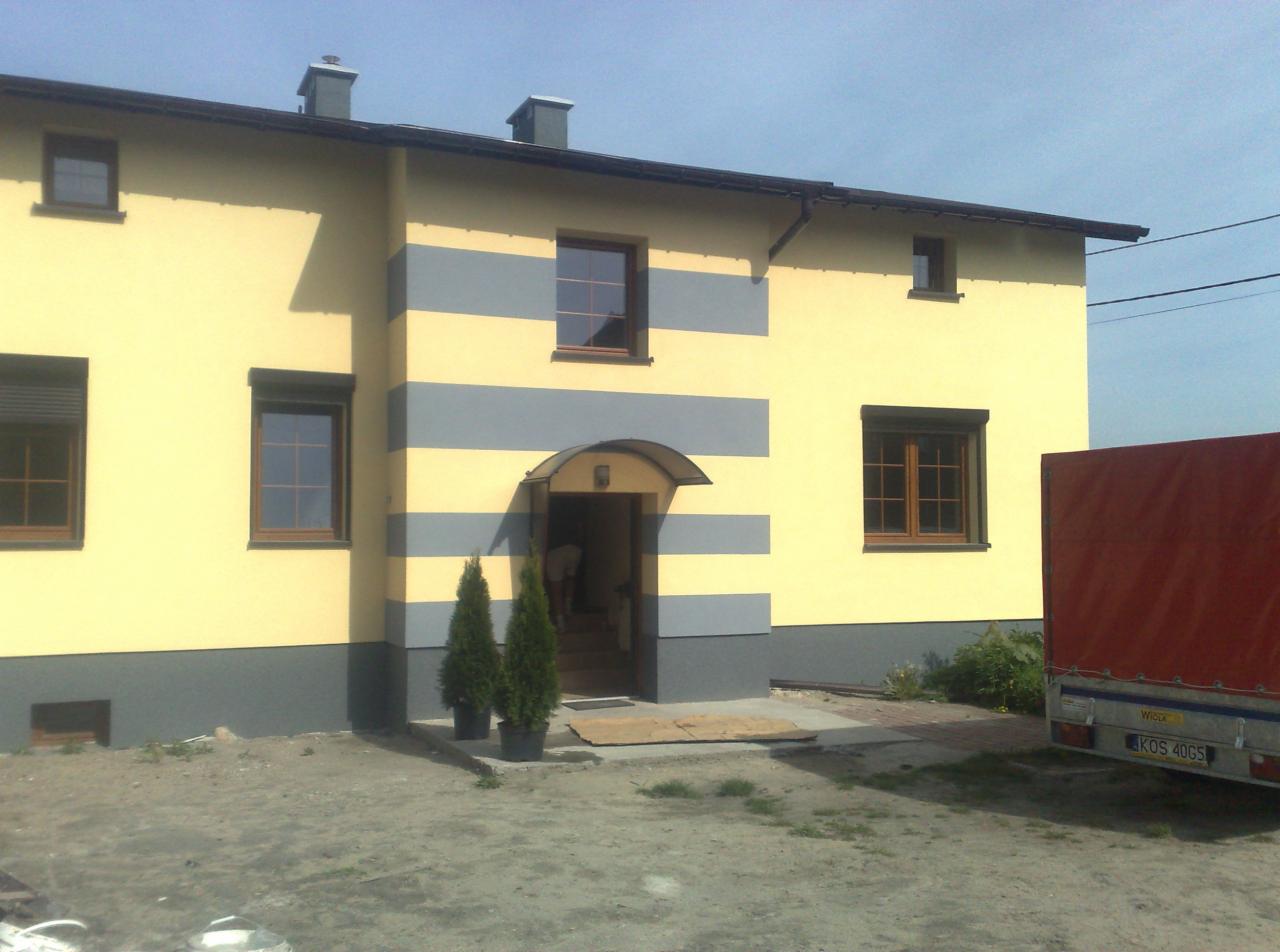 Dwukondygnacyjny dom z żółto-szarą elewacją w poziome pasy, widoczne dwie małe choinki w donicach przed wejściem i przyczepa samochodowa z czerwoną plandeką z boku budynku.