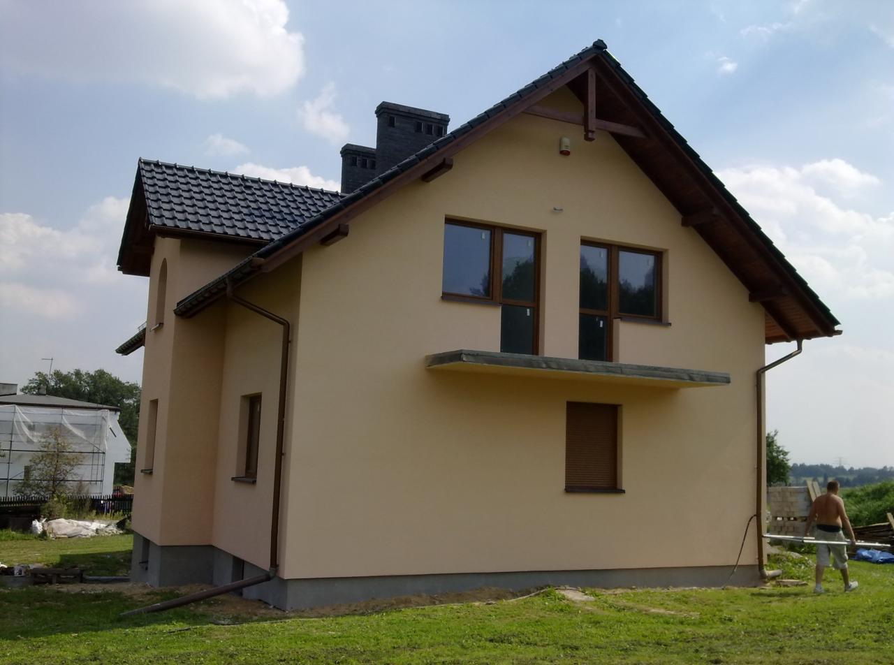 Gotowy dom jednorodzinny z beżową elewacją, ciemną dachówką i brązowymi oknami, widok z boku na tle zielonego trawnika i błękitnego nieba z chmurami, z widocznym mężczyzną przenoszącym rury.