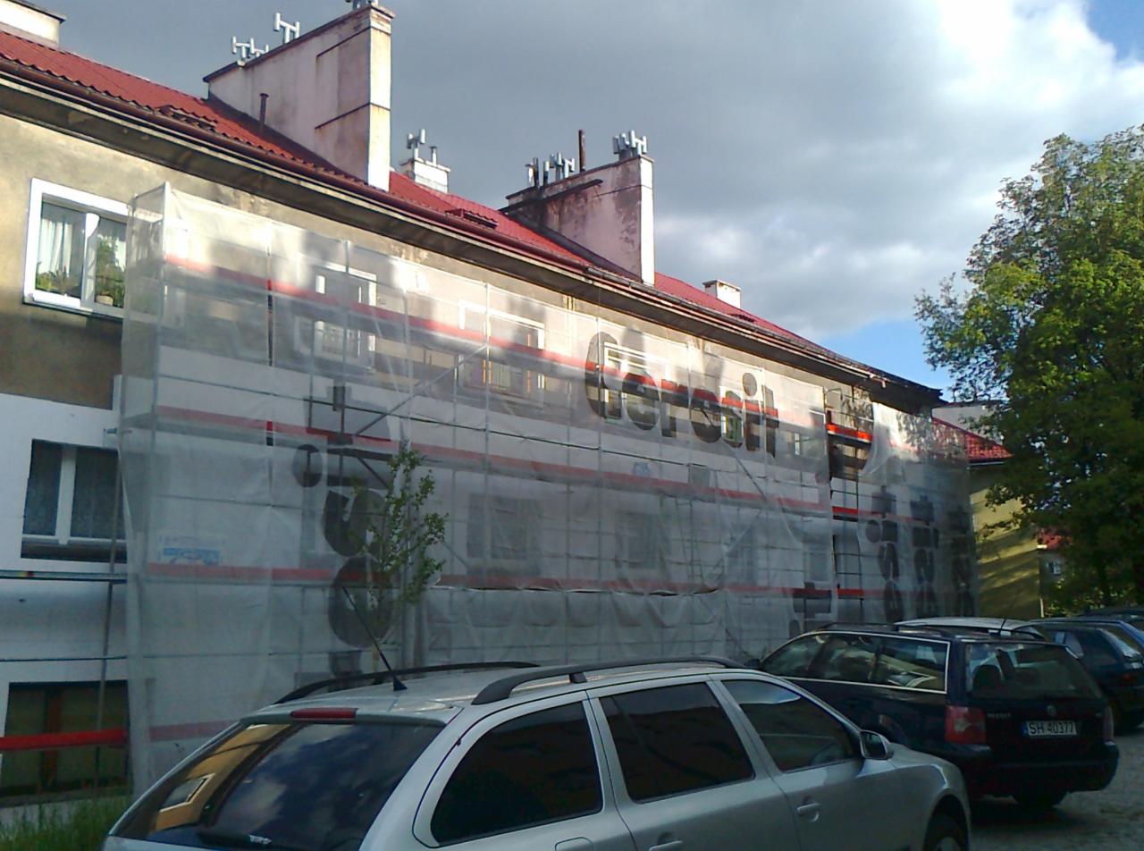Budynek mieszkalny w trakcie prac elewacyjnych, osłonięty siatką z nadrukowanym logo producenta materiałów budowlanych, widoczne rusztowania i zaparkowane samochody.