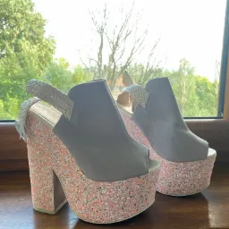 Para szarych butów na platformie z brokatem w odcieniach pomarańczy i bieli, stojących na drewnianym parapecie okiennym z widokiem na zieleń drzew.