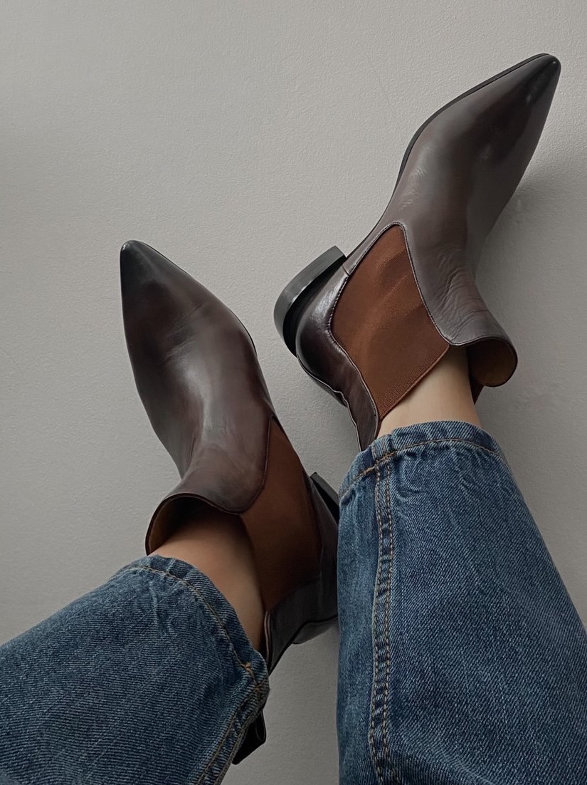 Para brązowych skórzanych botków chelsea z elastycznymi wstawkami, noszonych z dżinsami na tle szarej ściany.