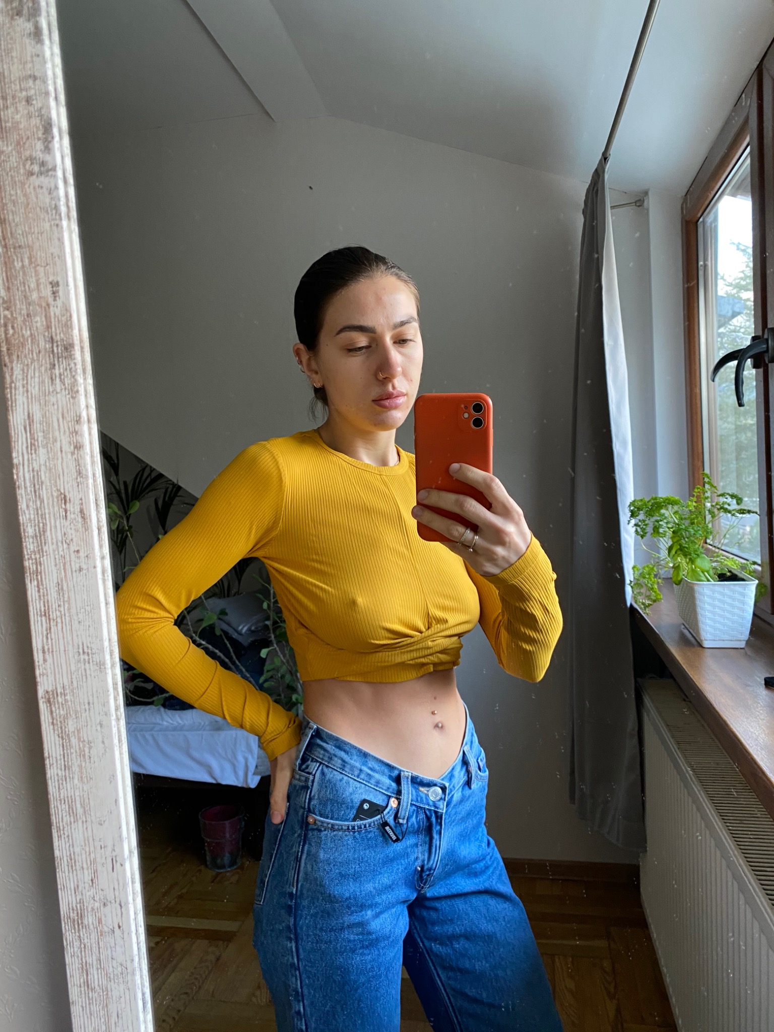 Osoba robiąca selfie w lustrze, ubrana w żółtą bluzkę z długim rękawem i szerokie jeansy, widoczny fragment sypialni z rośliną w tle.
