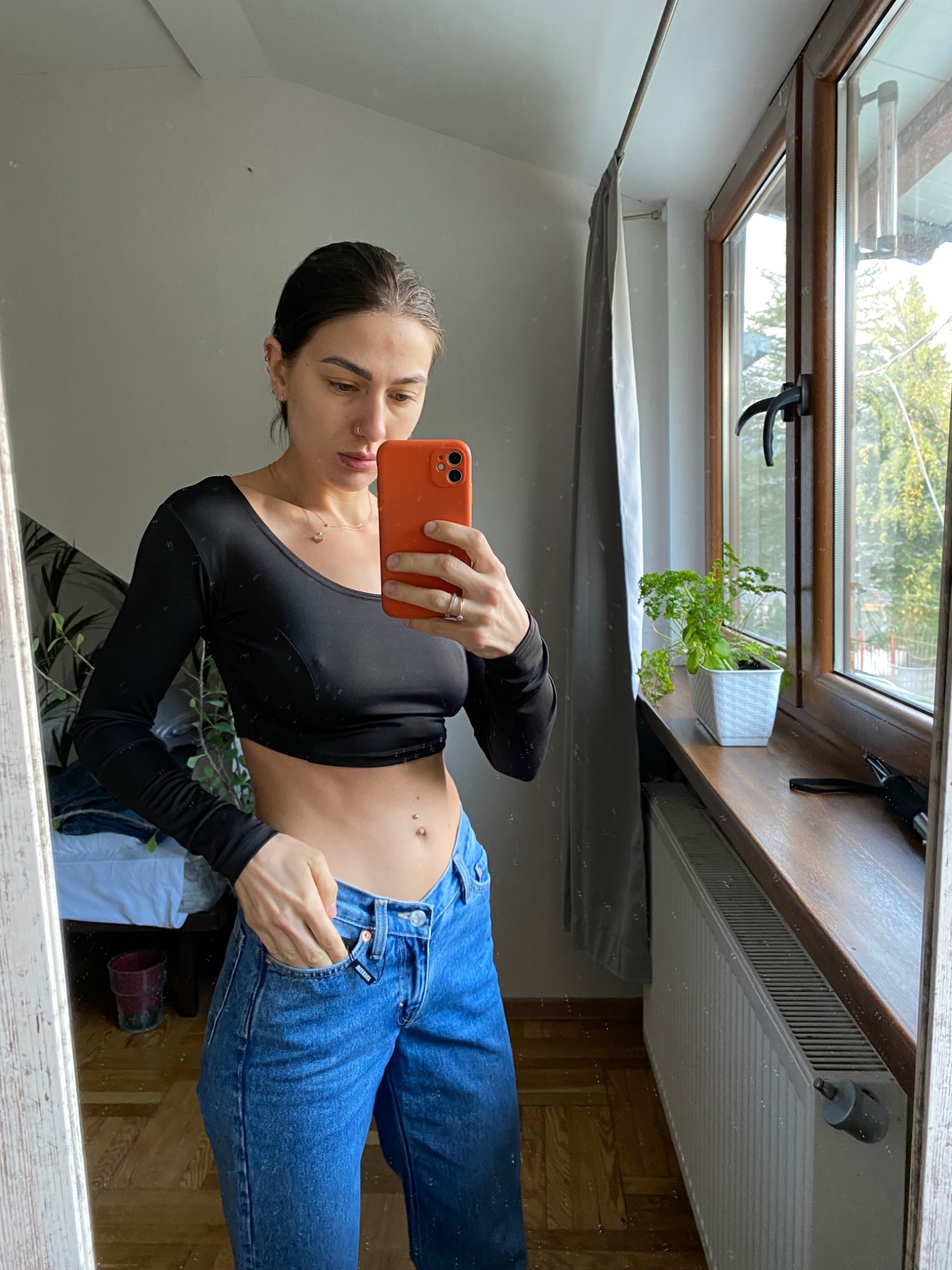 Osoba robiąca selfie w lustrze, ubrana w czarny crop top z długim rękawem i szerokie jeansy z ręką w kieszeni, na tle okna z rośliną doniczkową.