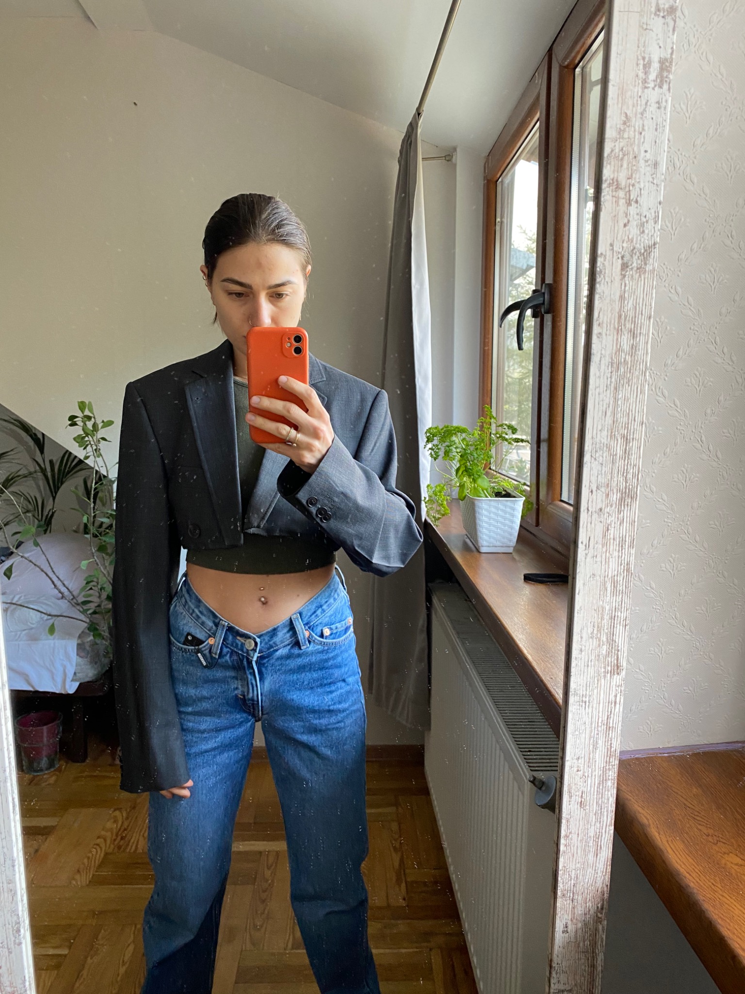 Osoba robi selfie w lustrze, ubrana w krótki czarny żakiet i dżinsy z niskim stanem, widoczny piercing w pępku, w tle okno z rośliną w doniczce i grzejnik.