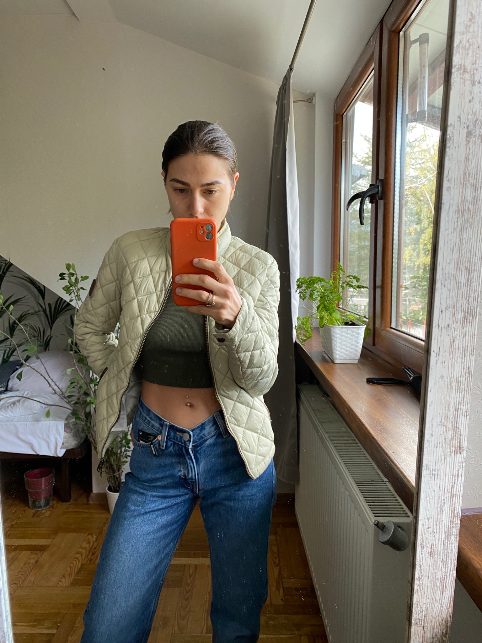 Osoba robi selfie w lustrze, ubrana w pikowaną kurtkę w odcieniu ecru, zielony top i szerokie jeansy z nisko osadzonym pasem, widoczne rośliny doniczkowe i okno w tle.