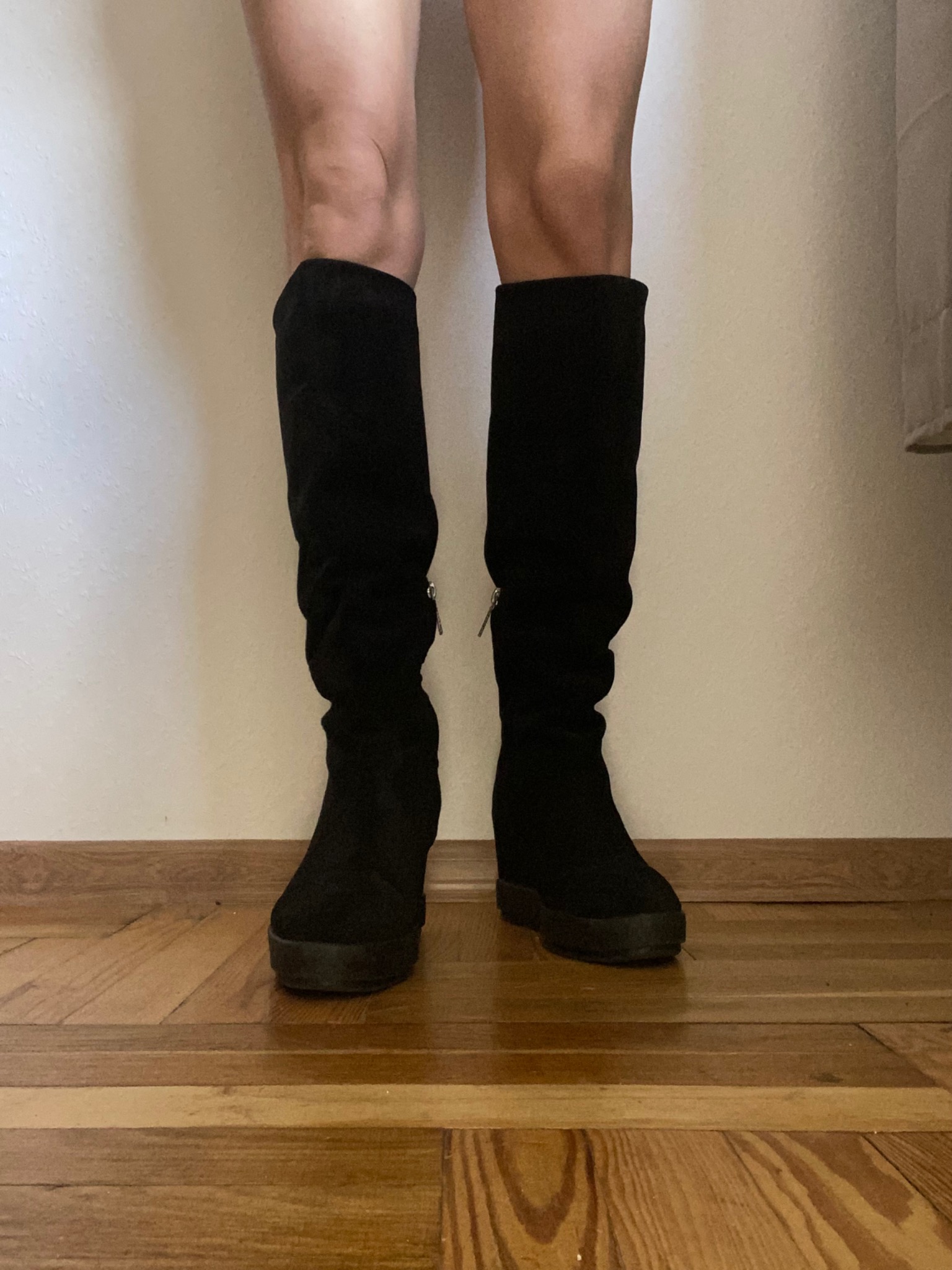 Osoba prezentuje czarne, zamszowe kozaki do kolan na niskiej platformie, z bocznym zamkiem błyskawicznym, stojąc na parkiecie z jasnego drewna.