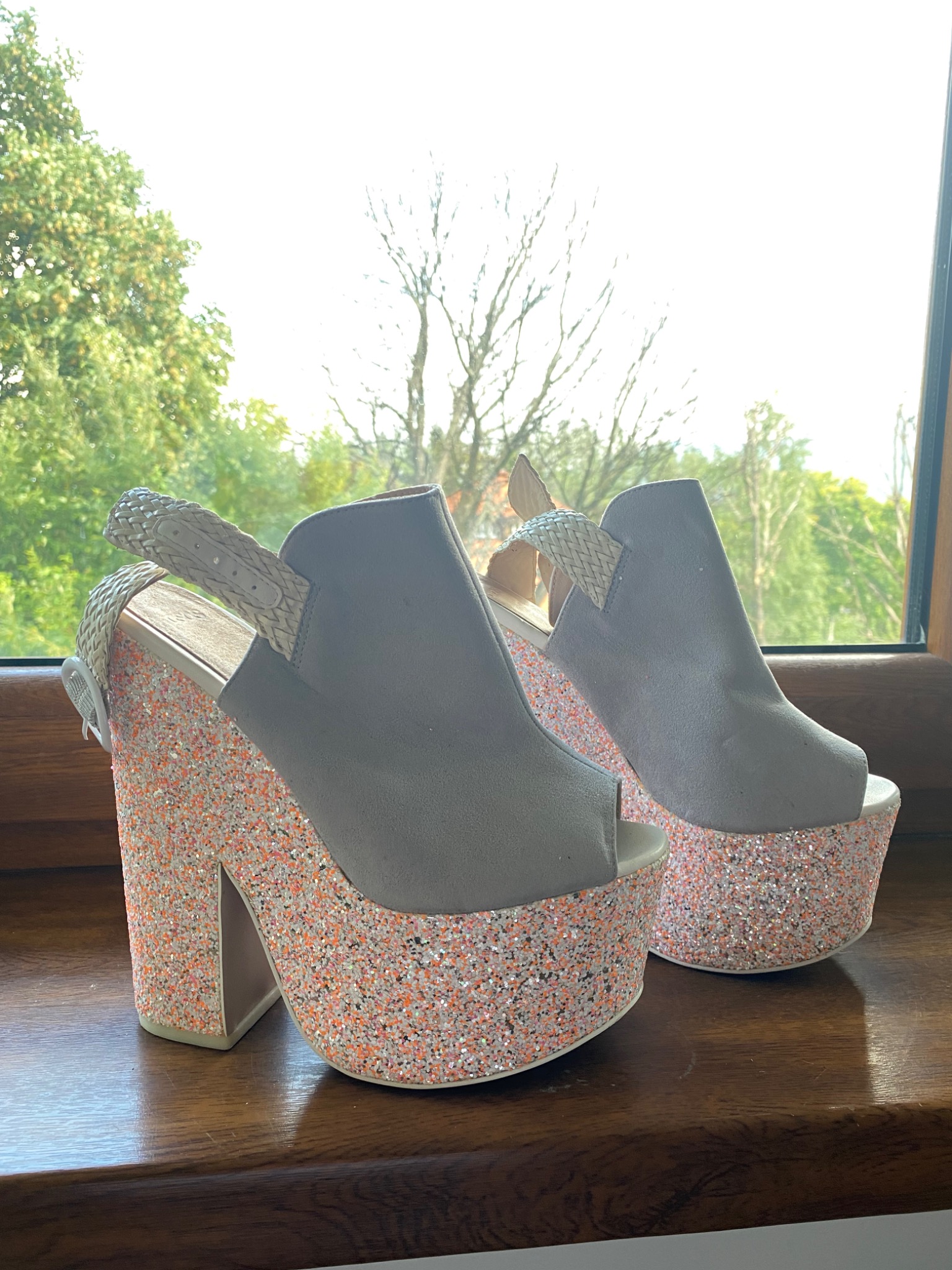 Para szarych butów na platformie z brokatem w odcieniach pomarańczy i bieli, stojących na drewnianym parapecie okiennym z widokiem na zieleń drzew.
