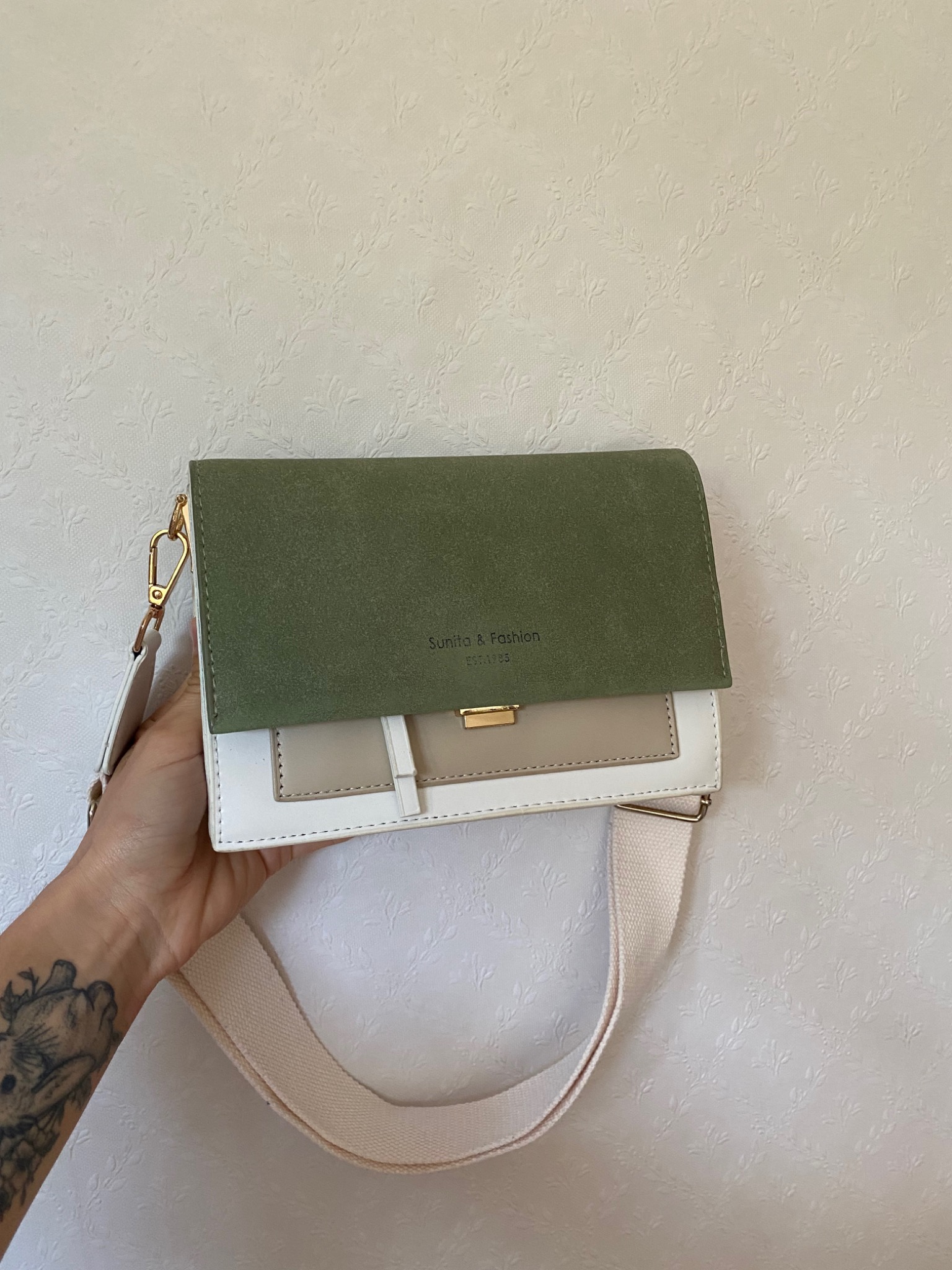 Trójkolorowa torebka listonoszka (oliwkowy, beżowy, biały) z szerokim paskiem na ramię, trzymana w dłoni z widocznym tatuażem na przedramieniu, na tle jasnej ściany z delikatnym wzorem.