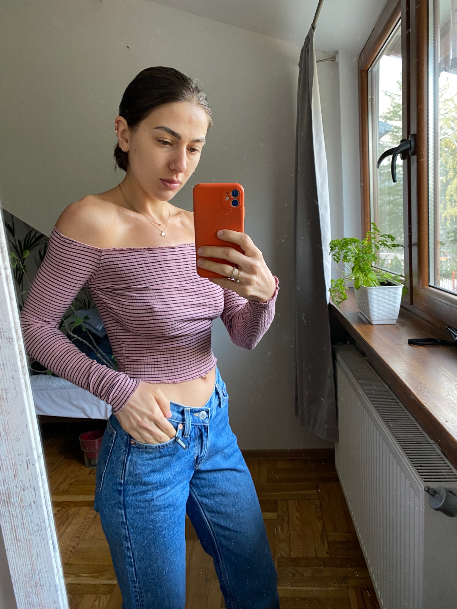 Osoba robiąca selfie w lustrze, ubrana w bluzkę w paski z odkrytymi ramionami i dżinsy, widoczny parapet z rośliną w tle.