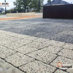 Wzornik z ekspozycją ułożonej kostki brukowej w trzech kolorach: jasnoszary, ciemnoszary i pomarańczowy, z logo Darbud w rogu, prezentowany na zewnątrz w punkcie sprzedaży Moszczanka.