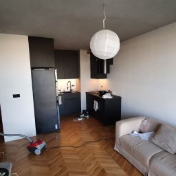 Miczada - Wnętrze mieszkania po remoncie, z widoczną częścią salonu z beżową sofą i aneksem kuchennym z czarnymi meblami, podłoga wyłożona parkietem w jodełkę, w tle widoczny odkurzacz.
