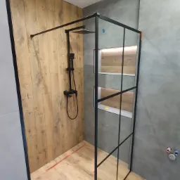 Nowoczesna kabina prysznicowa typu walk-in z czarnymi profilami, drewnopodobne płytki na ścianie i podłodze, szara ściana z wnękami oświetlonymi LED.