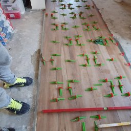 Miczada - Układanie płytek imitujących drewno z użyciem poziomicy i klipsów dystansowych; widoczne buty robocze i butelka coli