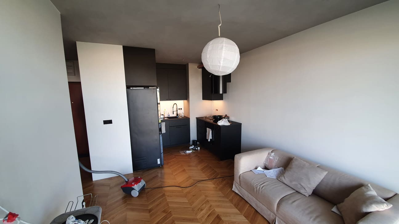 Wnętrze mieszkania po remoncie, z widoczną częścią salonu z beżową sofą i aneksem kuchennym z czarnymi meblami, podłoga wyłożona parkietem w jodełkę, w tle widoczny odkurzacz.