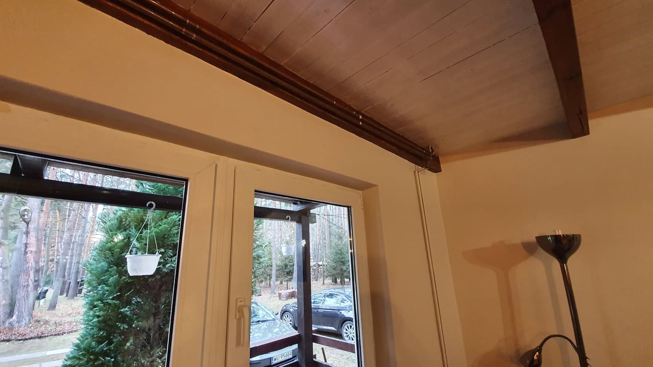 Wnętrze z drewnianym sufitem i oknem wychodzącym na ogród, widoczna lampa stojąca z ciemnym abażurem.